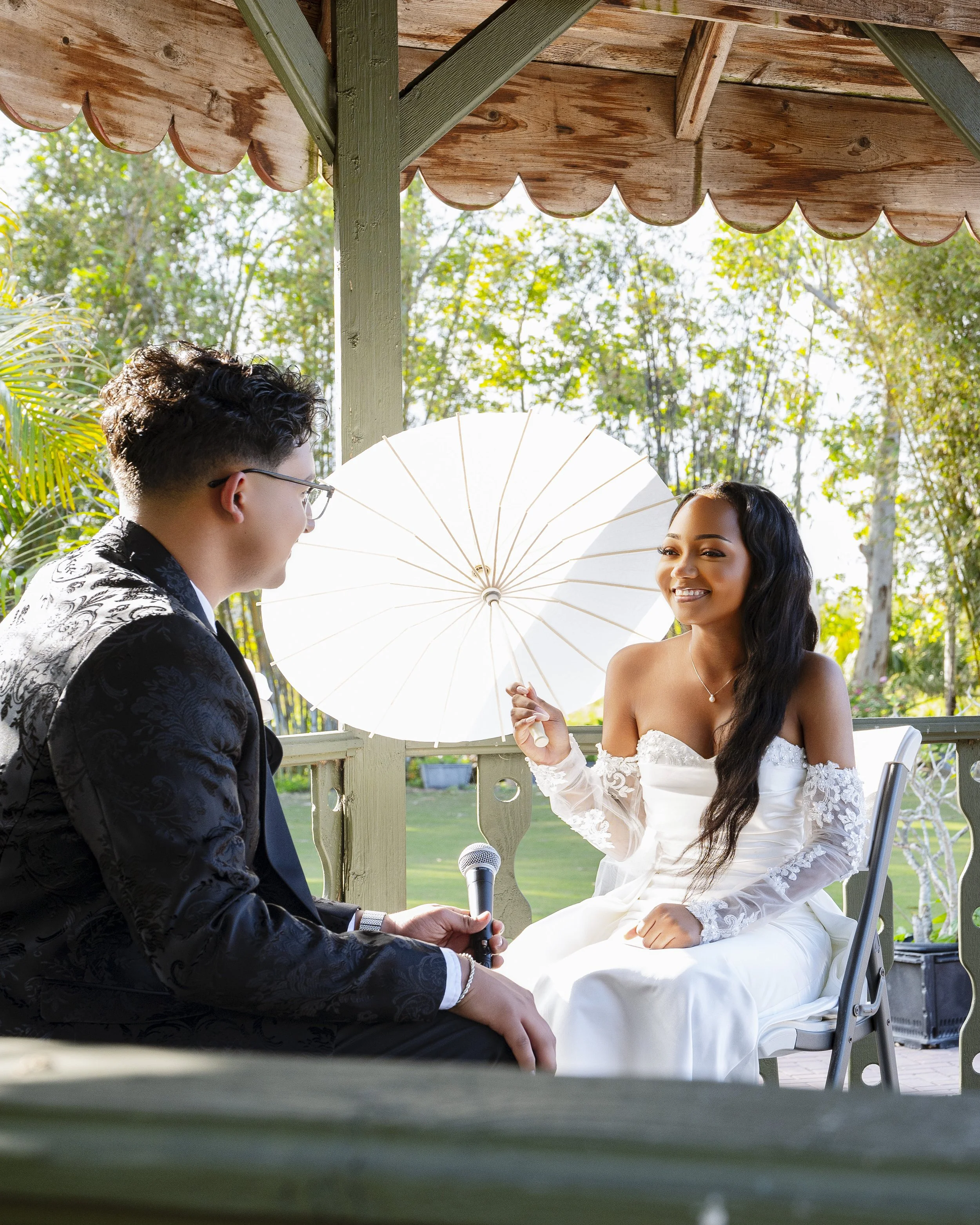 A woman in a white wedding dress holding an umbrella and smiling, sitting on a chair, while a man in a black suit and glasses, holding a microphone, is sitting nearby. They are outside on a porch with trees in the background.