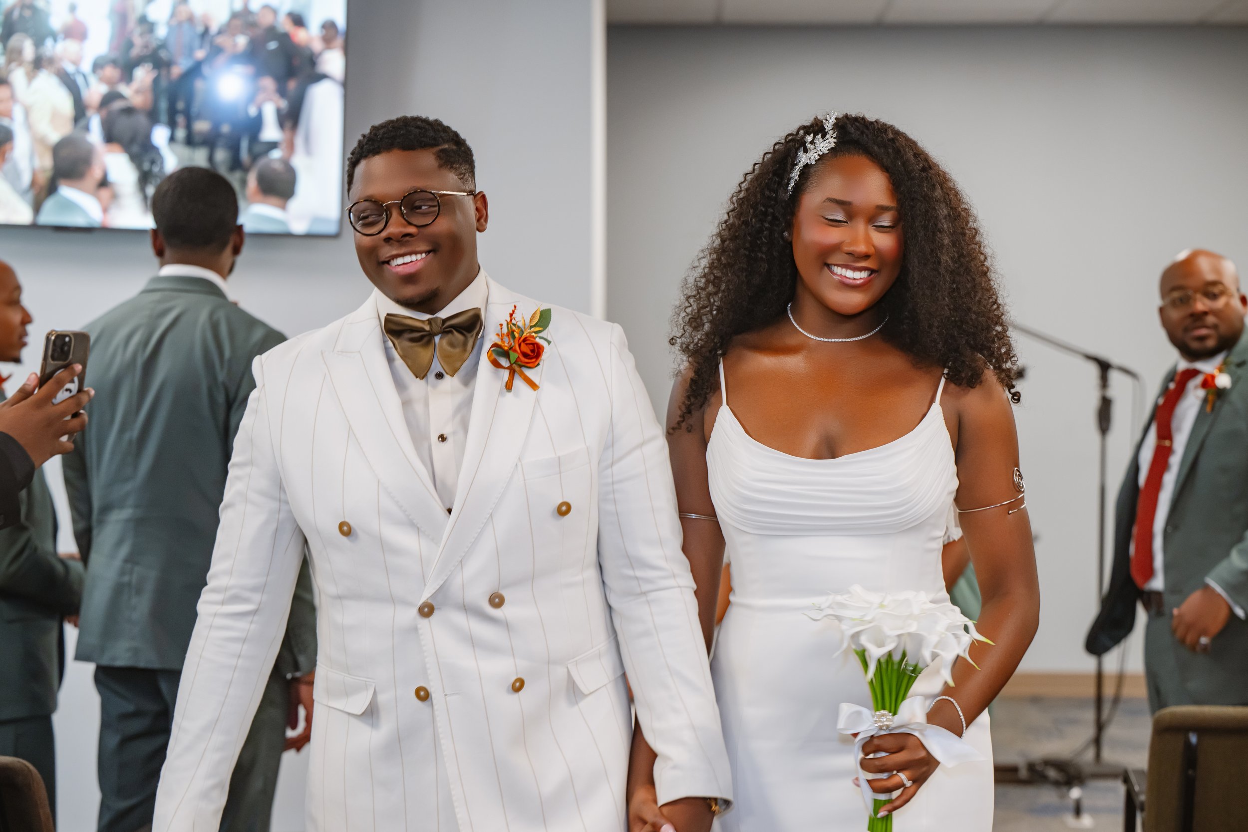 A couple, a man and a woman, celebrating their wedding in a church, dressed in white wedding attire, smiling, with the woman holding a bouquet of white calla lilies.