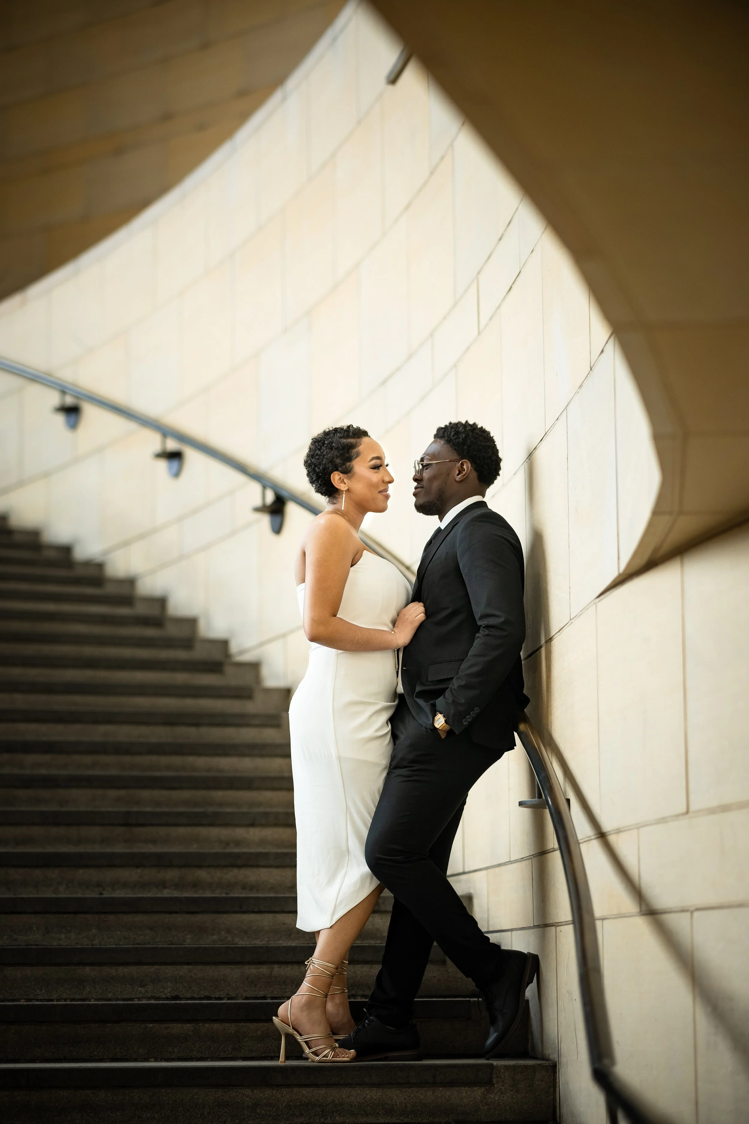 A stylish couple dressed in formal attire, standing on a staircase indoors, sharing an intimate moment.