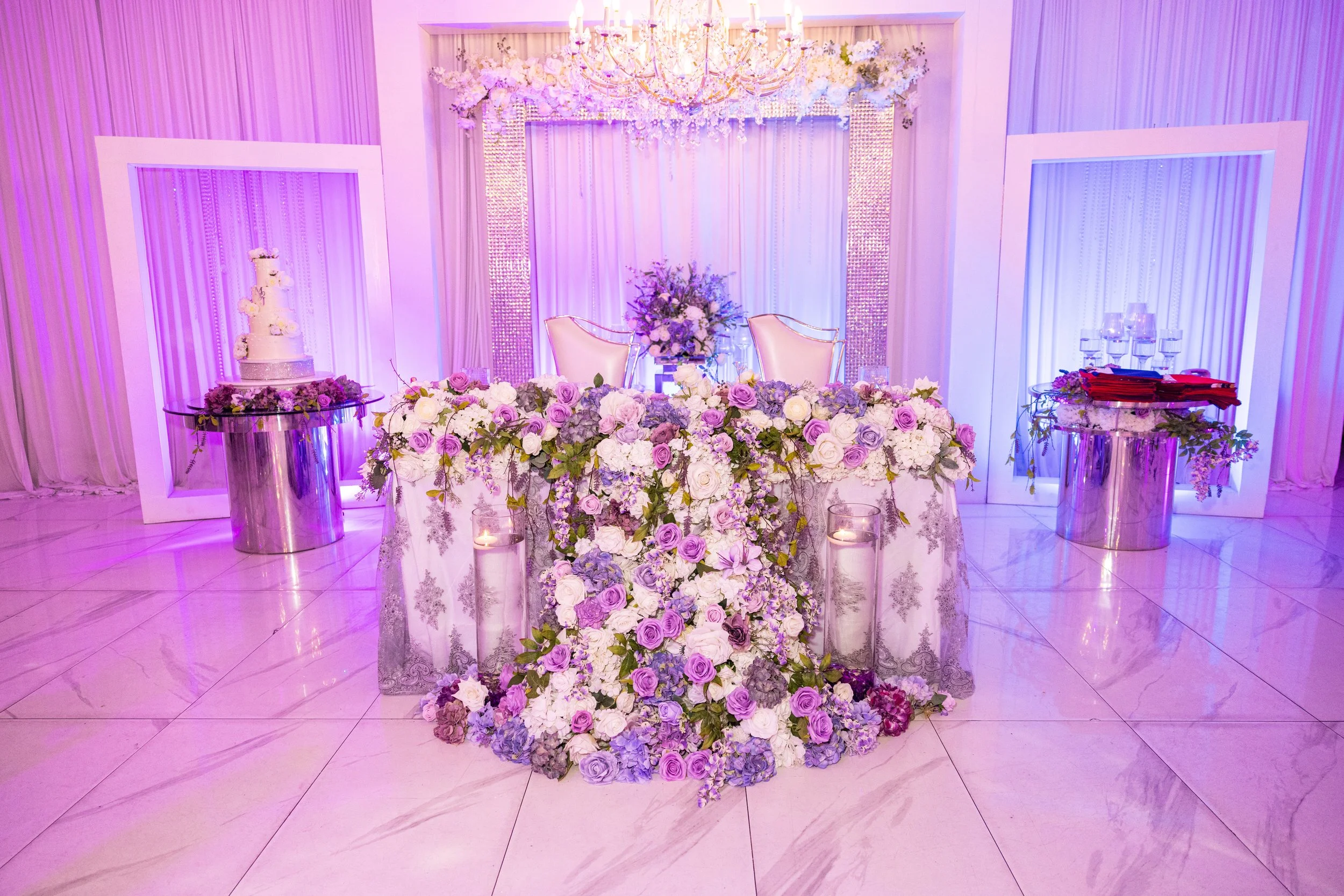 A wedding or reception stage decorated with purple and white flowers and drapes, featuring a chandelier, a tiered wedding cake, and floral arrangements on shiny silver tables.