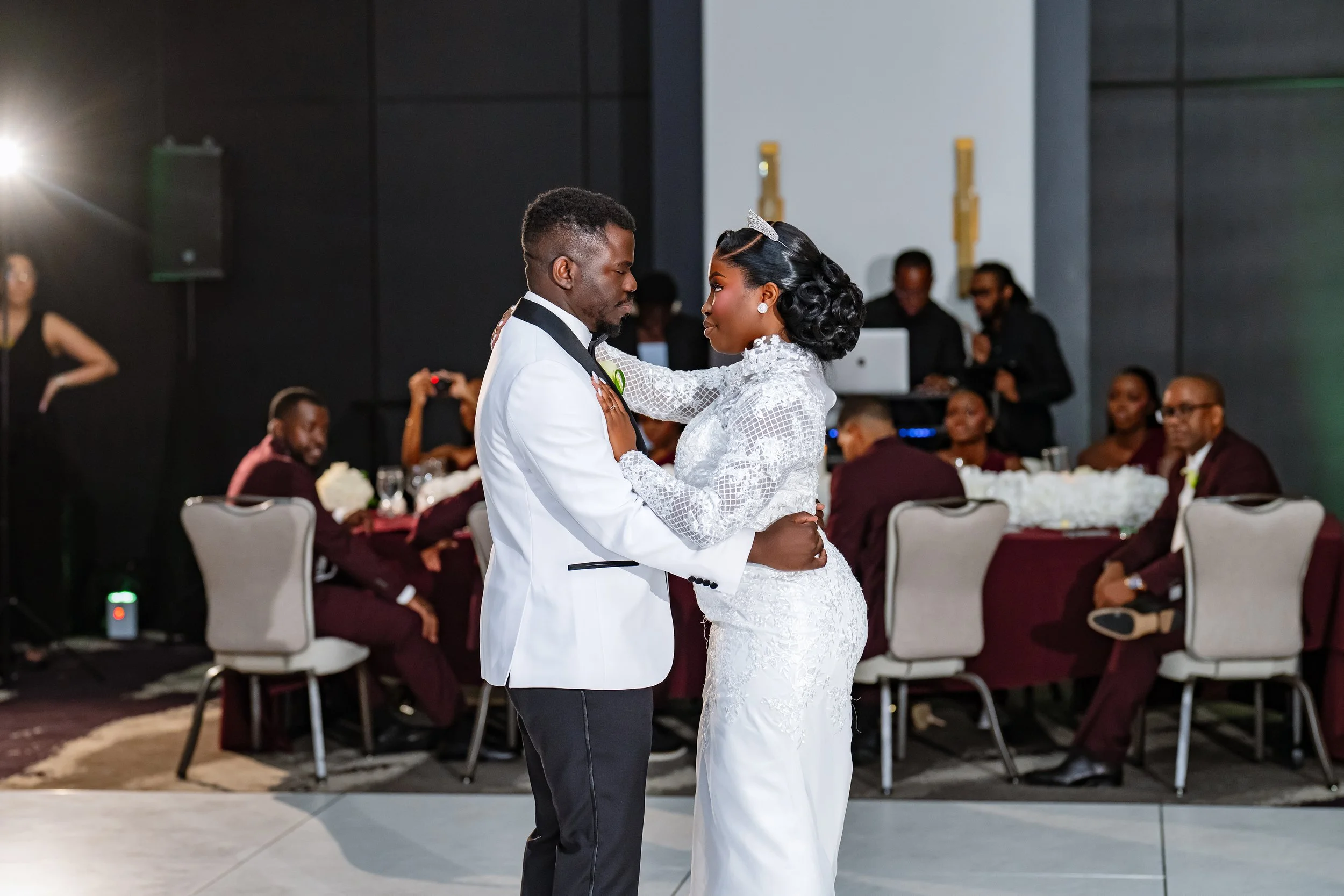 A couple dancing at their wedding reception, surrounded by seated guests in a dimly lit venue.