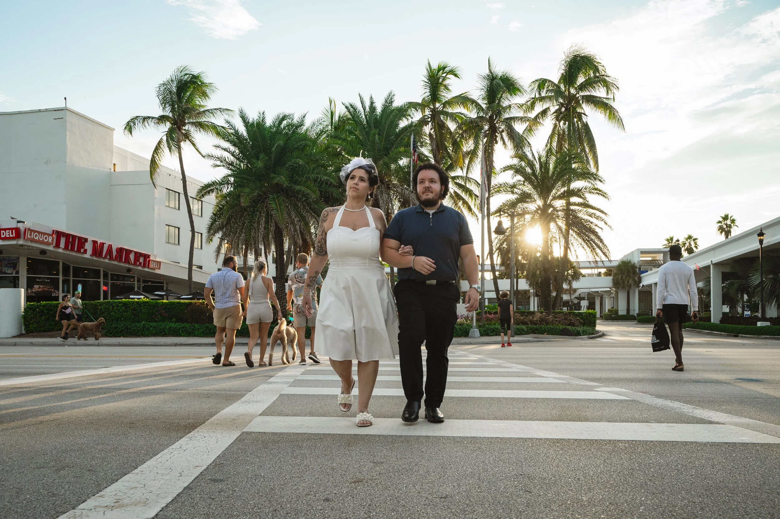 A couple walking across a crosswalk in a sunny urban area with palm trees and a strip mall in the background, while other pedestrians walk nearby.