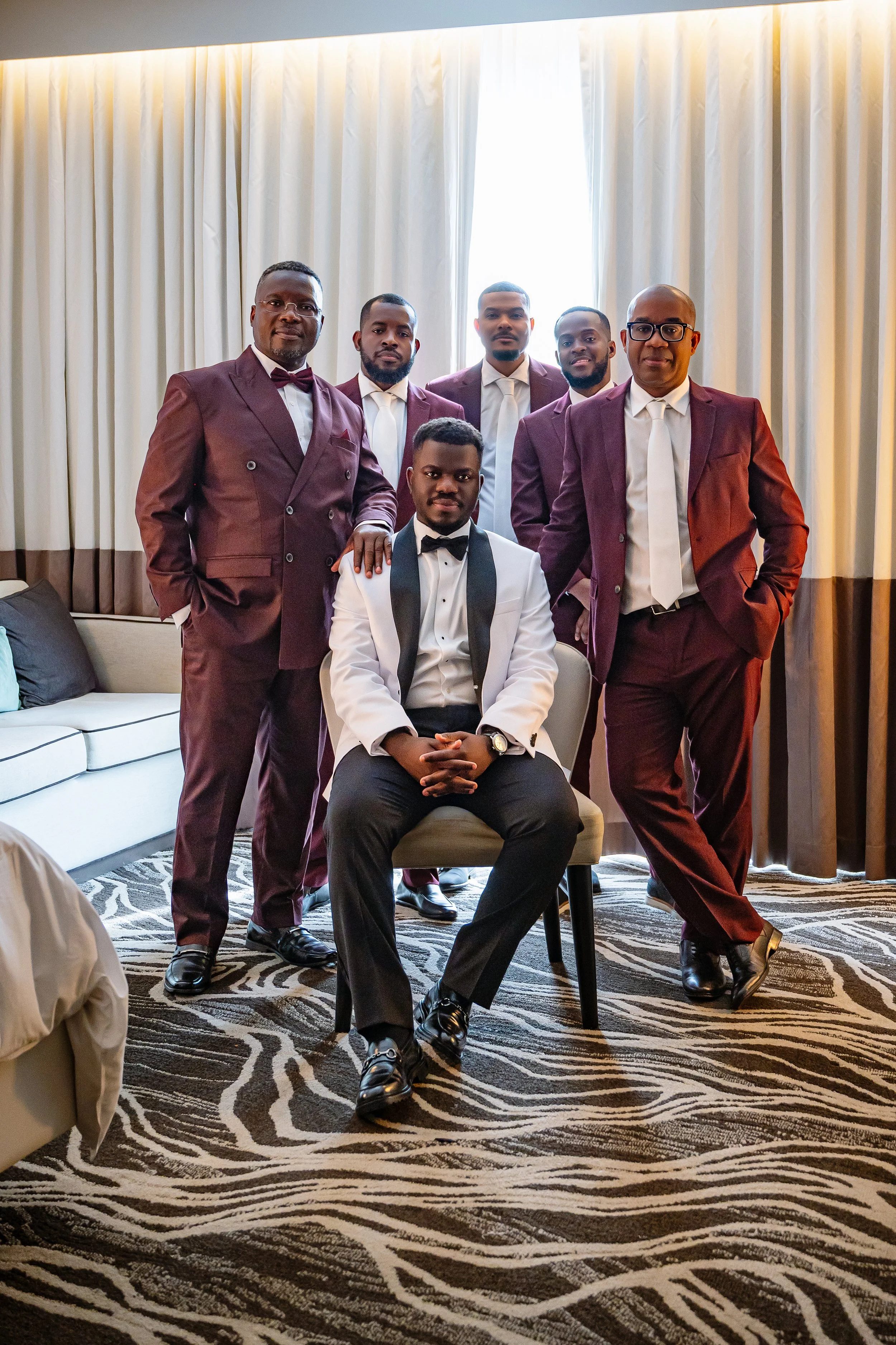 Seven men in formal suits posing for a photo in a room with curtains and a patterned rug, with one seated in front and the others standing behind.