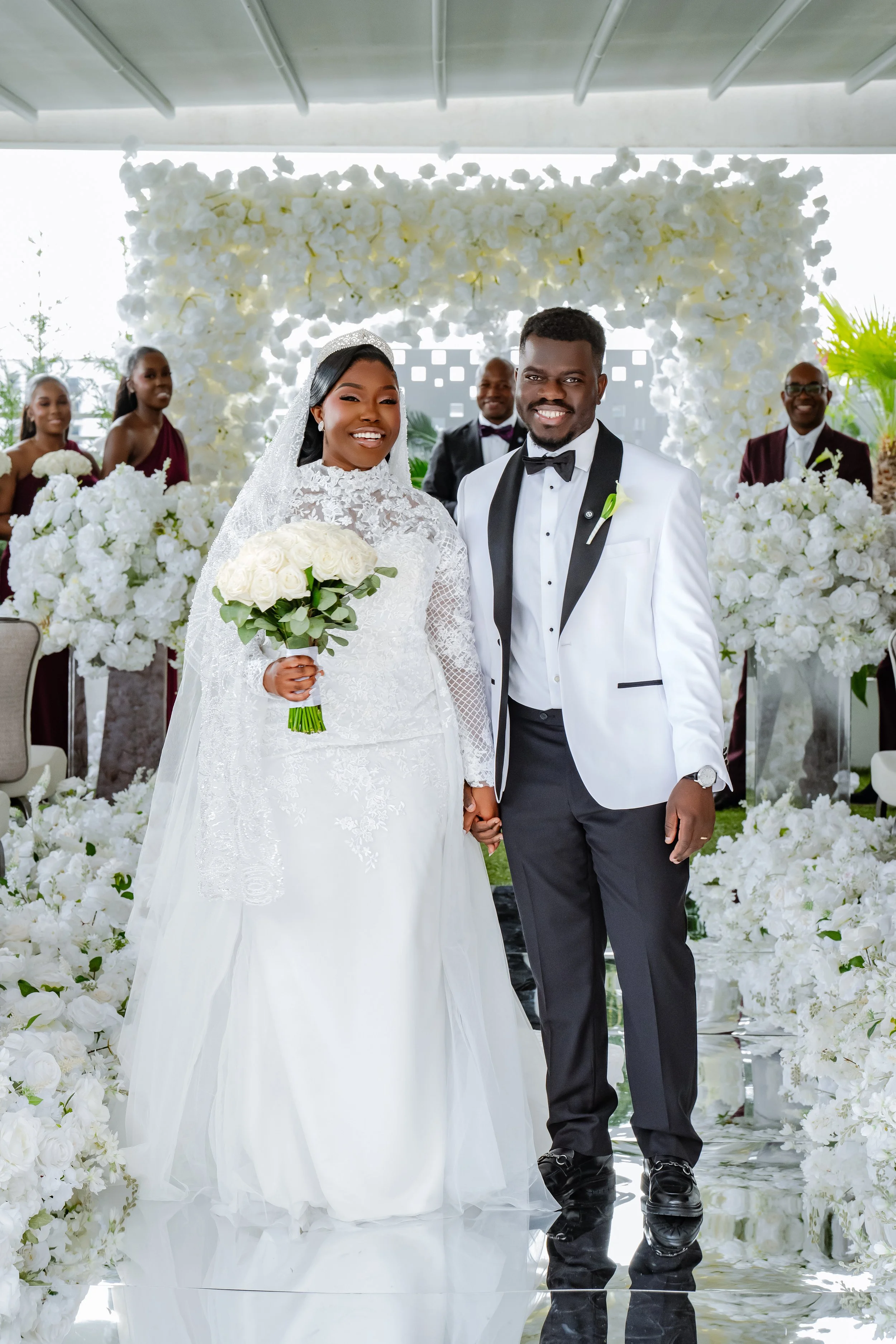 A bride and groom standing together at their wedding ceremony, surrounded by white floral decorations, with bridesmaids and groomsmen in the background.