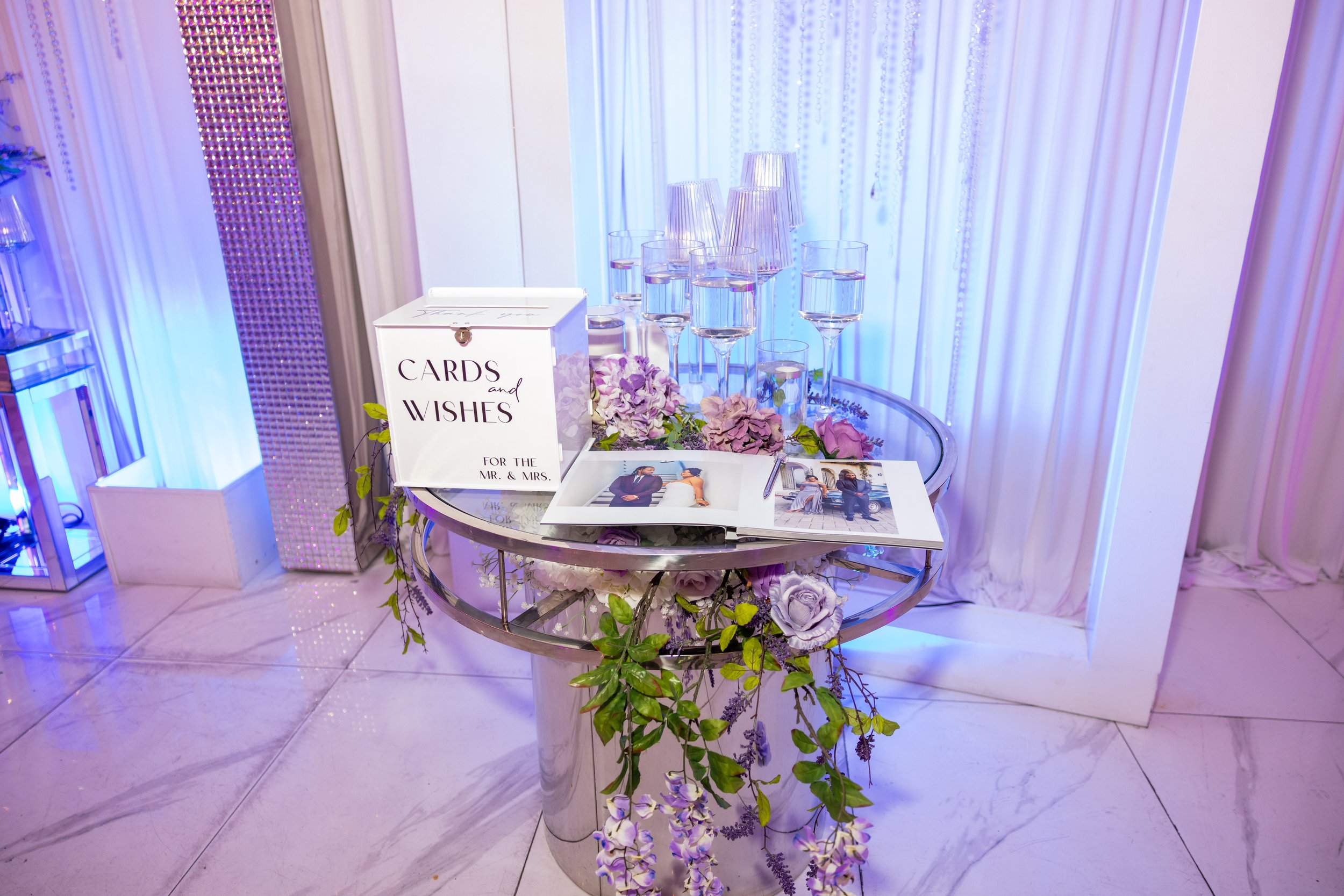Table with wedding cards, flowers, and drinks, set against a backdrop of white curtains with hanging beaded decorations, in a decorated event venue.