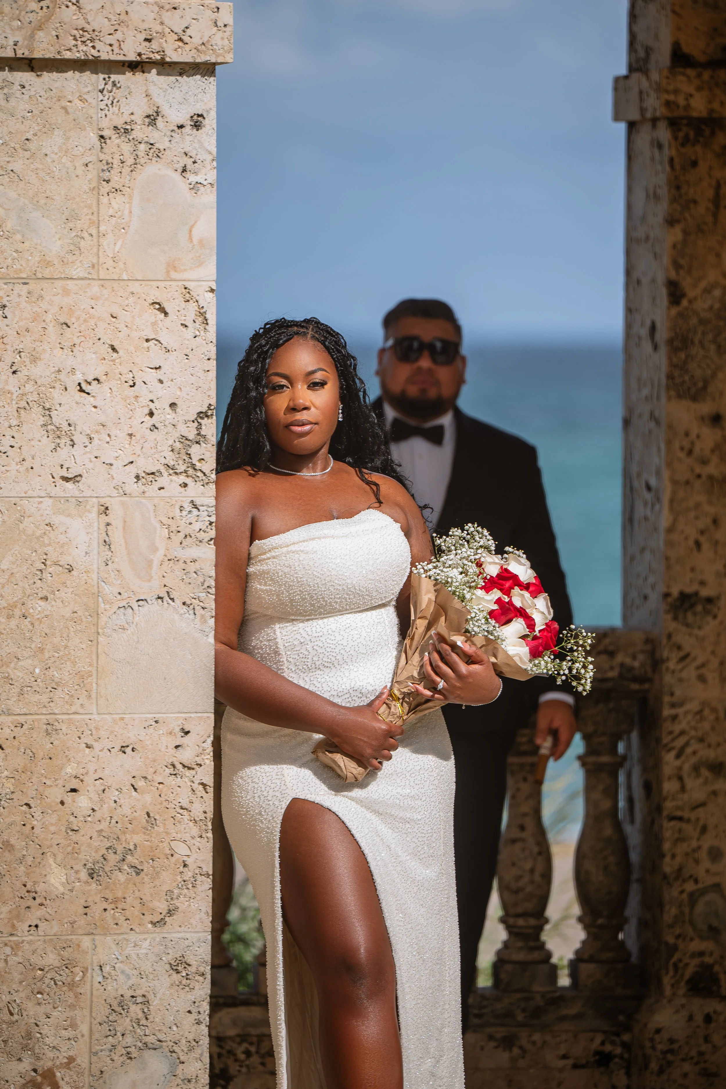 A bride holding a bouquet of flowers stands in front of a stone archway with a man dressed in a tuxedo in the background, overlooking the ocean.