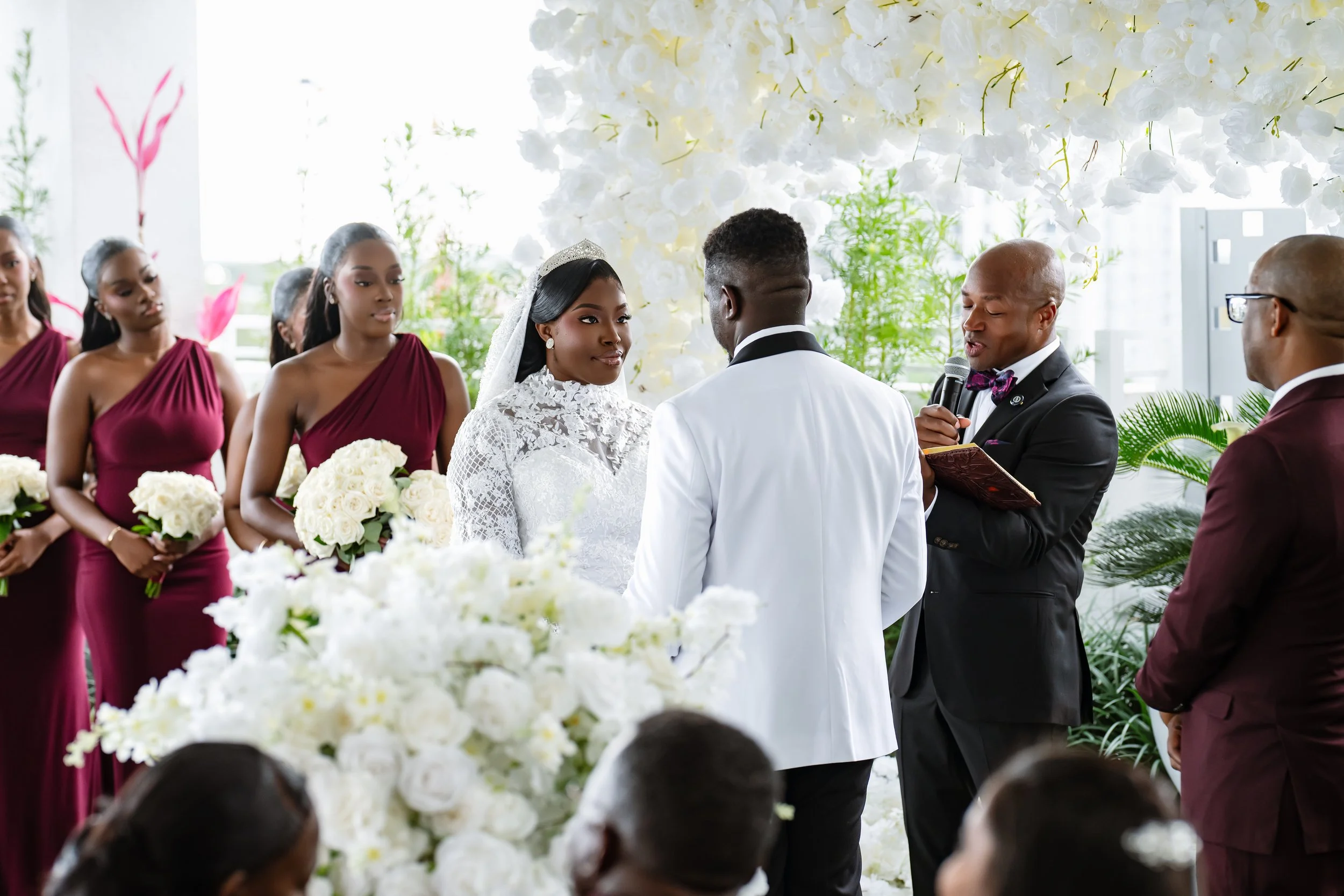 A wedding ceremony with a bride in white lace dress and veil, groom in white suit, and officiant in black tuxedo with bow tie, surrounded by bridesmaids in maroon dresses and floral arrangements, in a decorated venue with white flowers and green plan