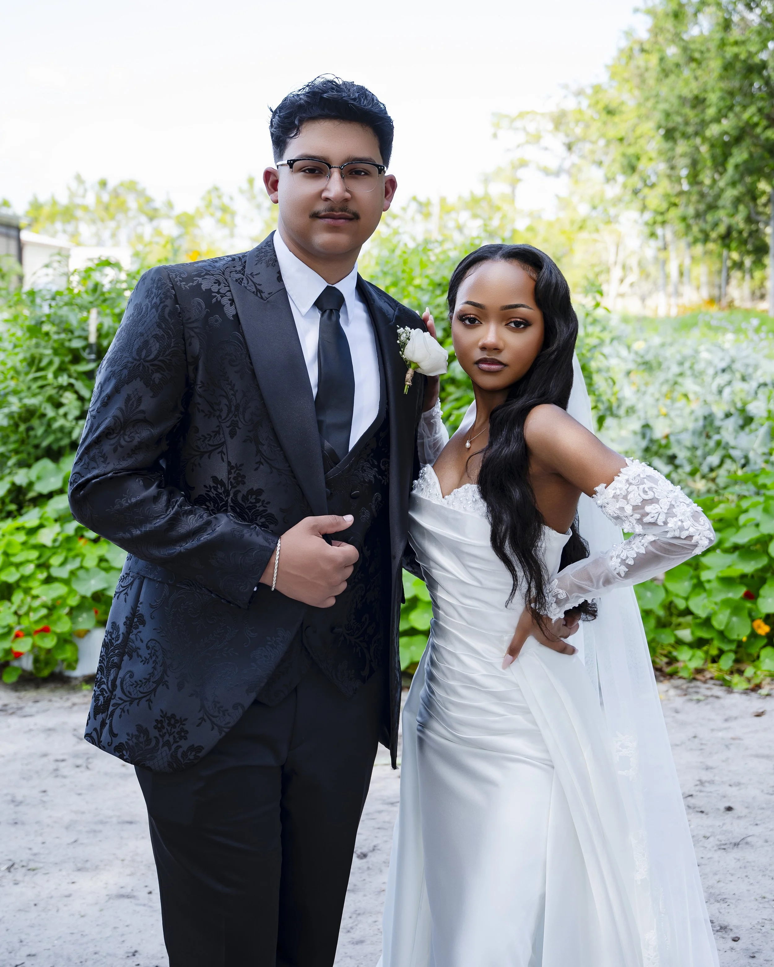 A bride and groom in wedding attire standing outdoors in front of green foliage.