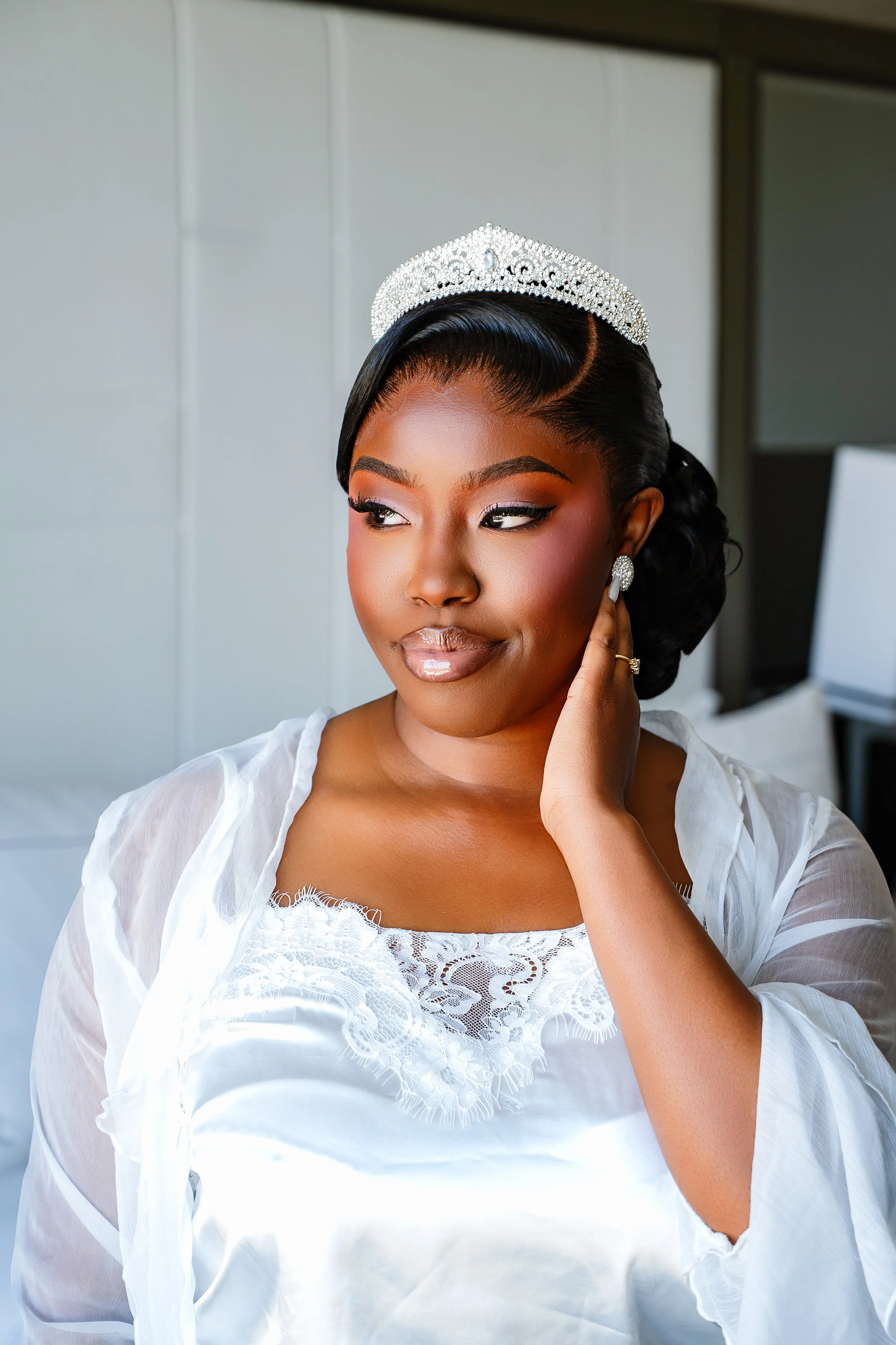 A woman with dark, styled hair wearing a tiara, earrings, and a white embroidered dress, touching her earring while looking away.