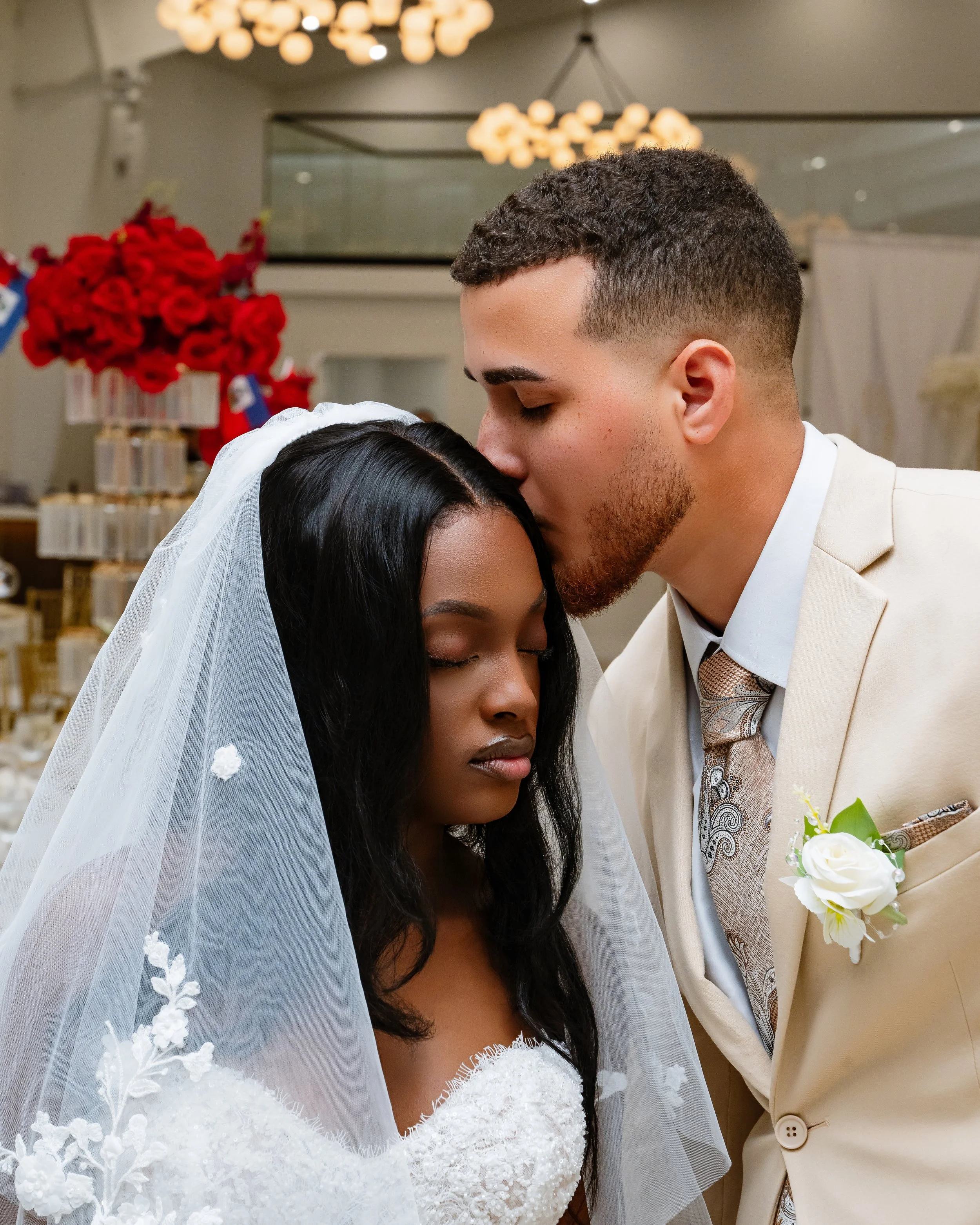 A groom kisses a bride on her forehead during a wedding ceremony in an elegant hall decorated with flowers and lights.