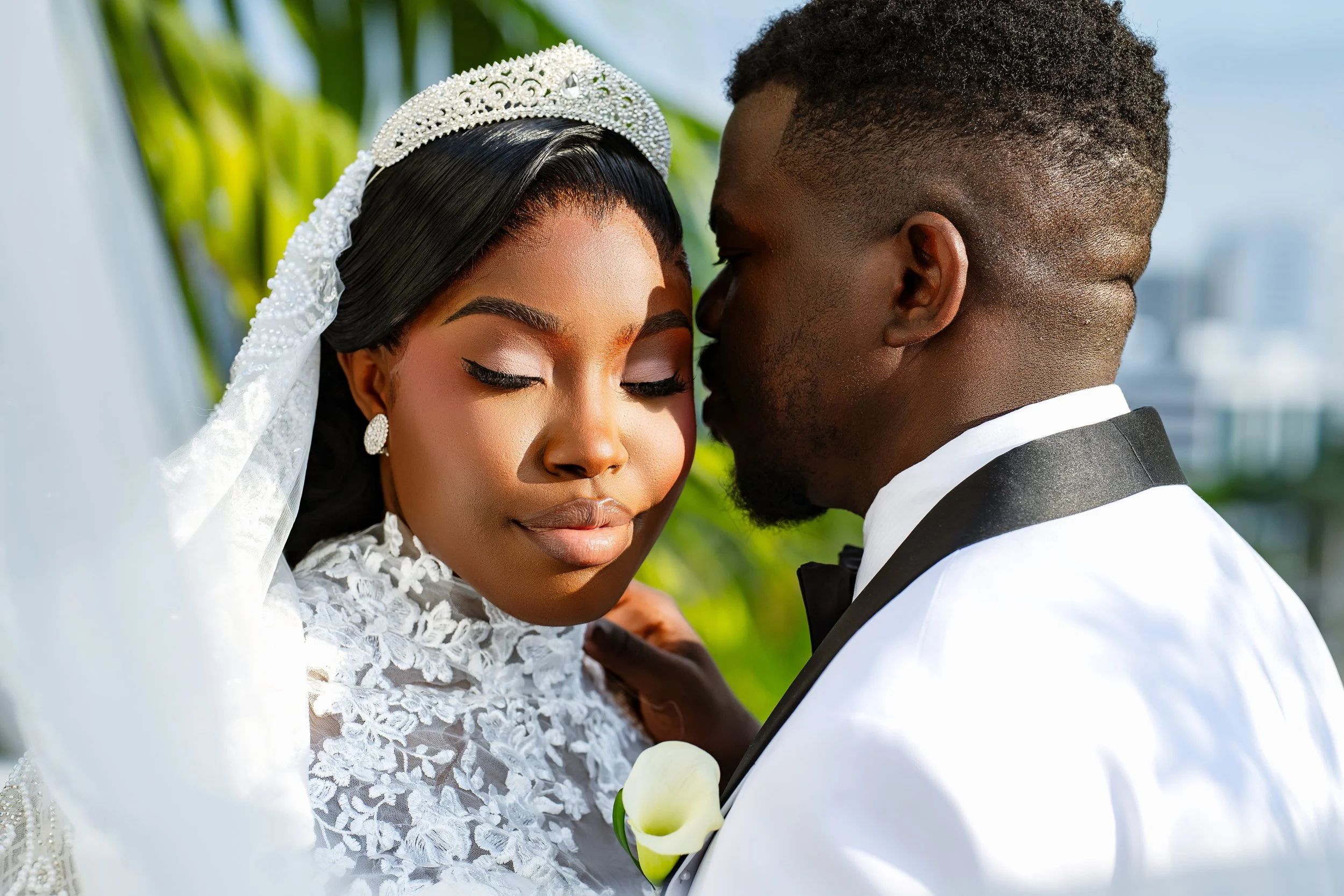 Close-up of a bride and groom on their wedding day, facing each other with eyes closed, outdoors with blurred greenery in the background.