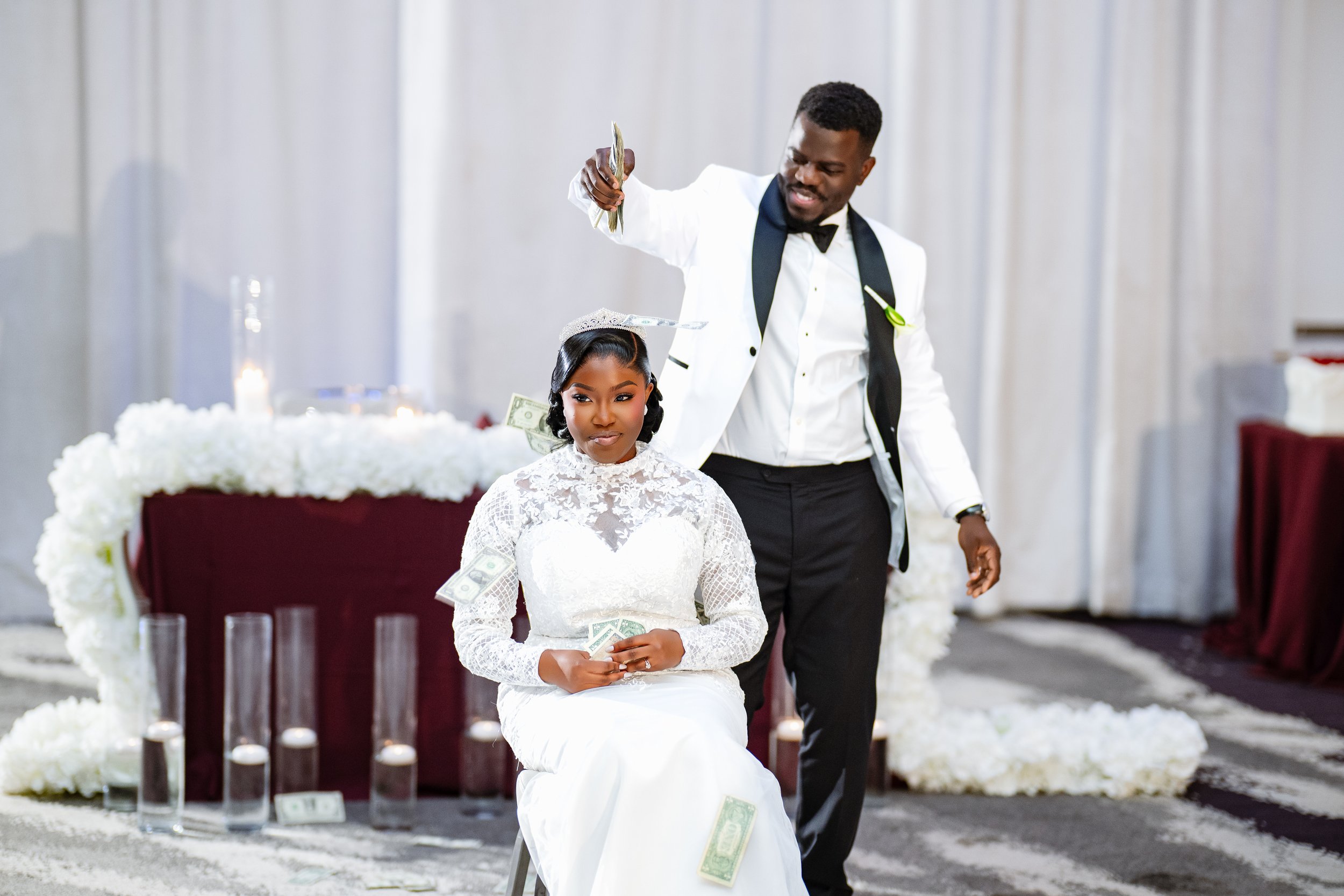 A bride in a white lace wedding dress and tiara sits holding dollar bills in her hands during a wedding ceremony, while a man in a white tuxedo and black bow tie stands behind her, throwing dollar bills in the air. The background features a decorated