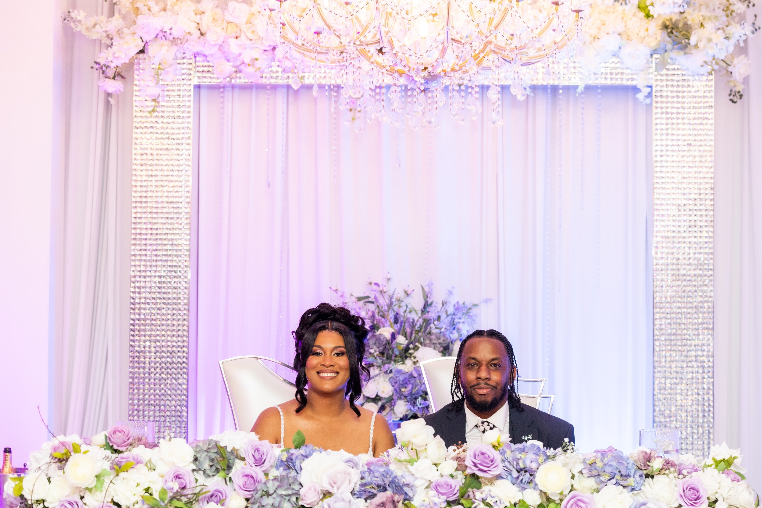A smiling woman in a wedding dress and a man in a suit sit at a decorated table with purple and white flowers, in front of a floral backdrop and a chandelier.