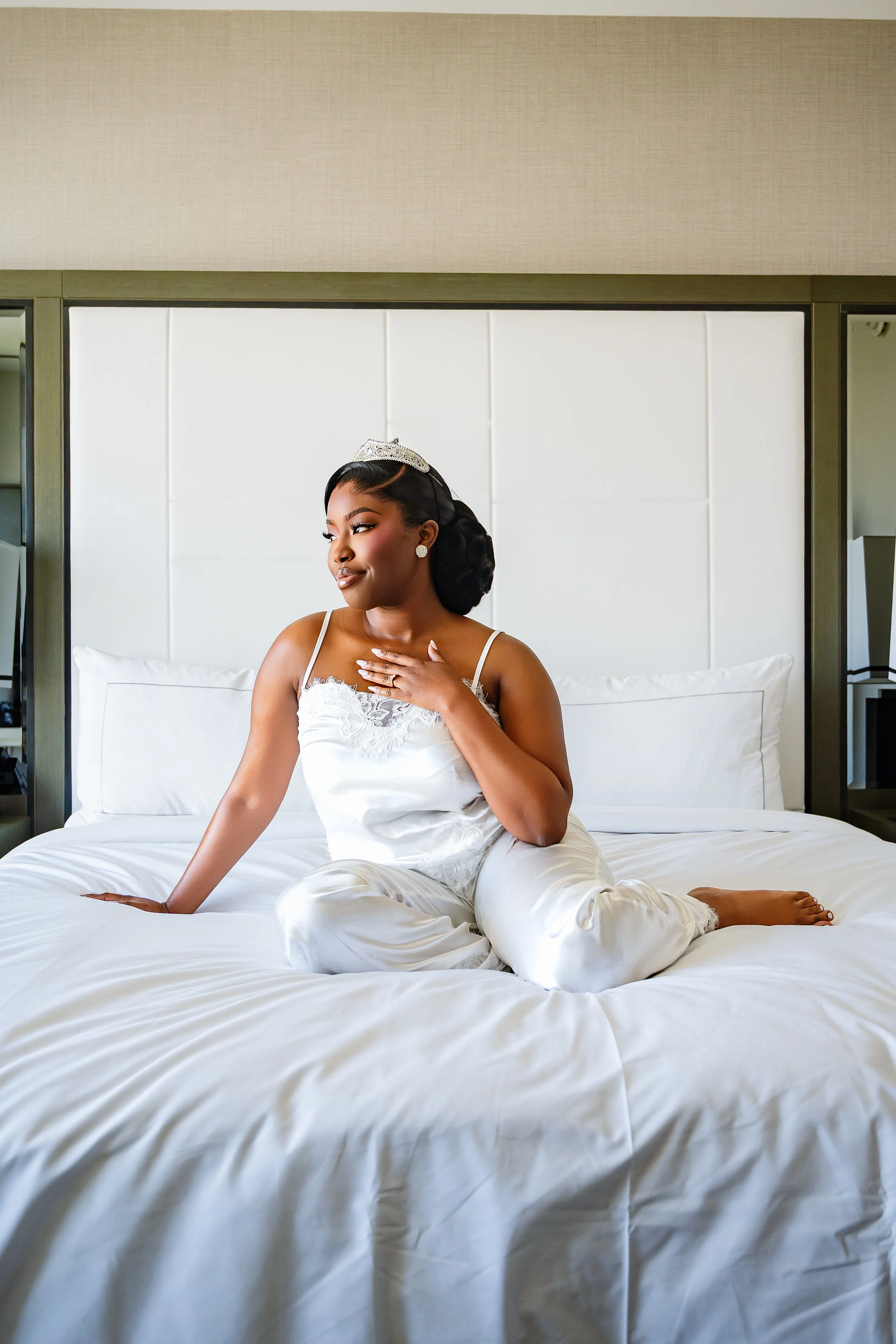 A woman in white satin pajamas sitting cross-legged on a bed with white sheets, resting one hand on the bed and the other on her chest, wearing a tiara and earrings, with a calm and content expression.