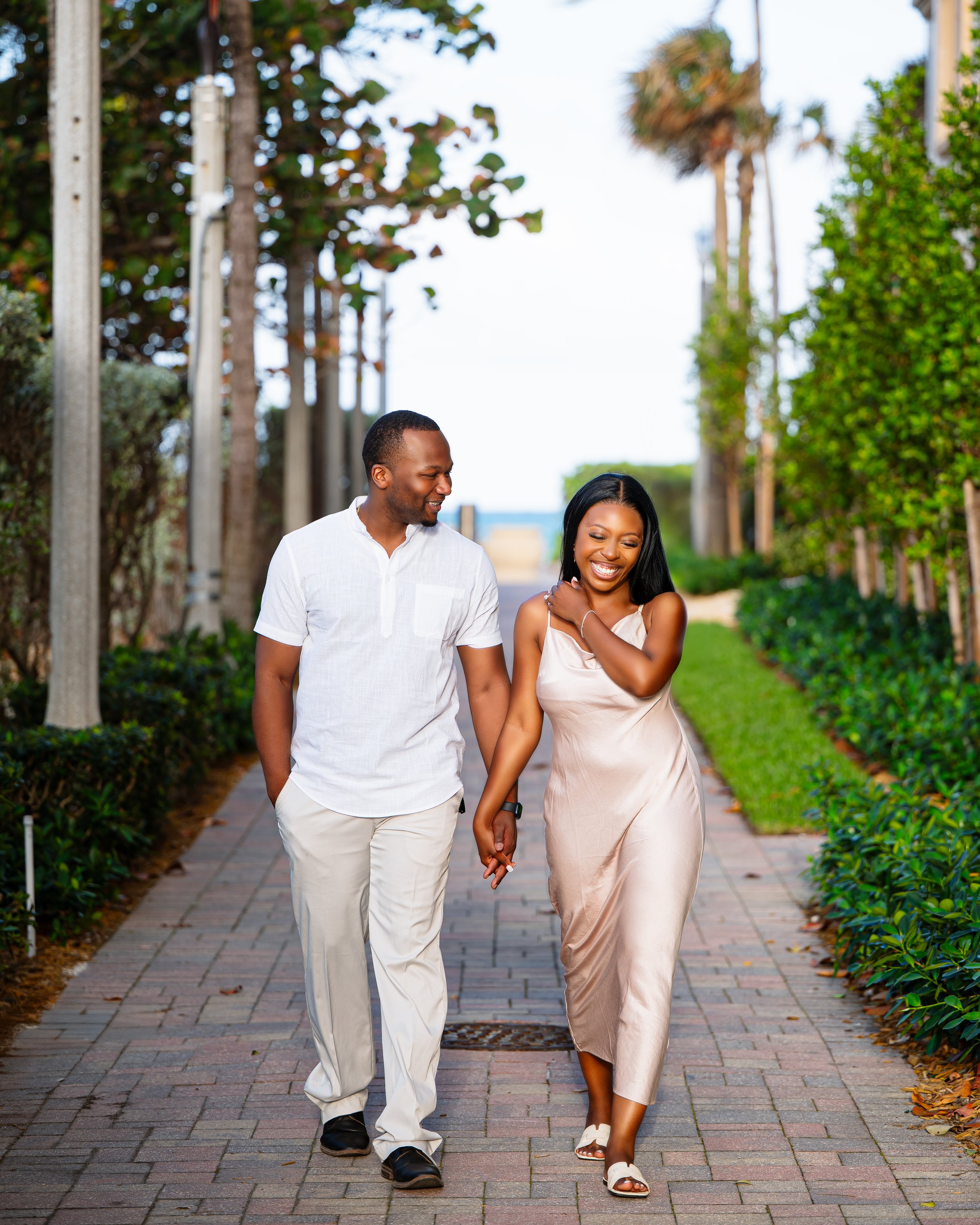 A smiling couple walking together on a brick sidewalk lined with green bushes and trees, holding hands and enjoying a sunny day.