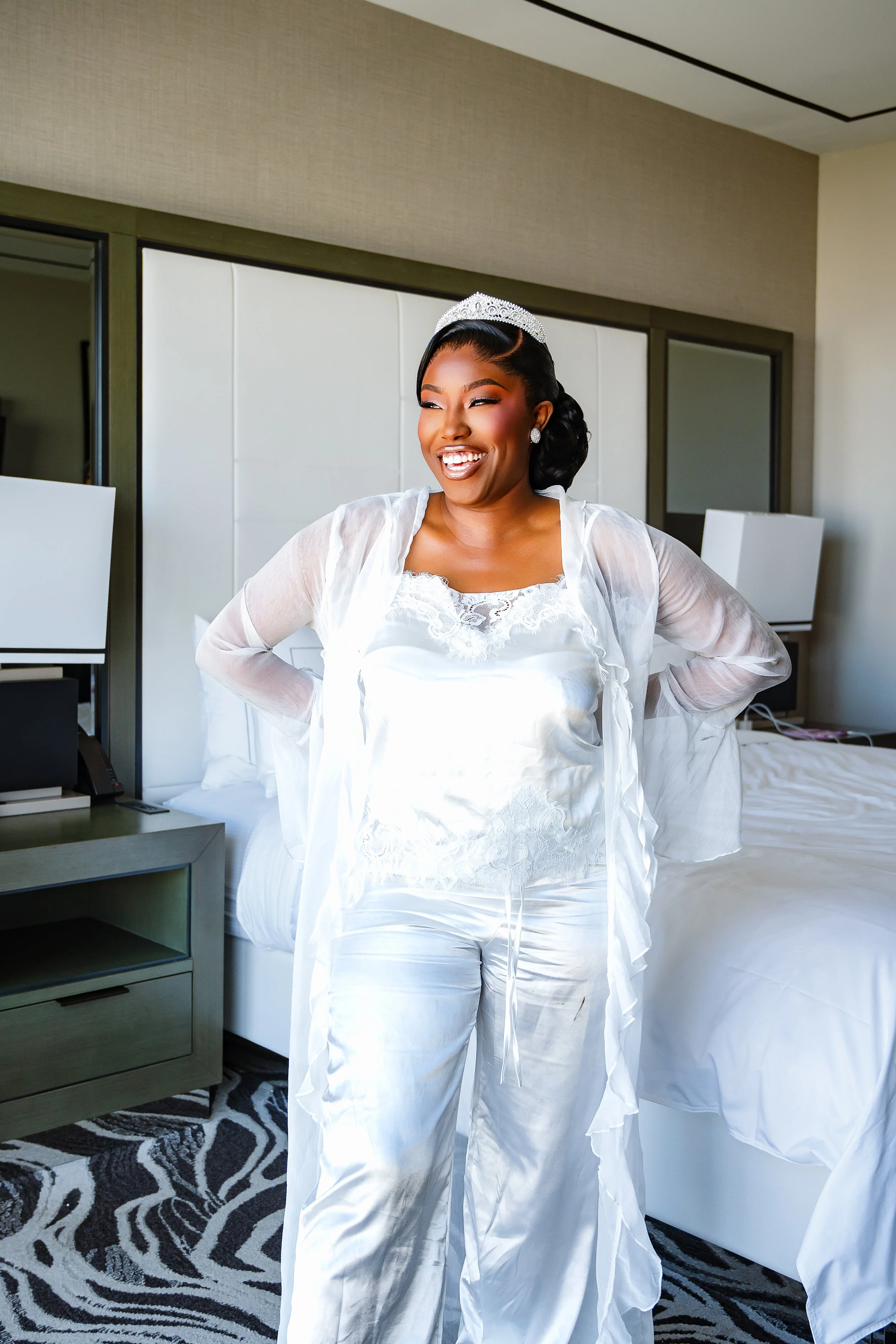 A woman dressed in white satin pajamas, wearing a tiara and earrings, smiling and posing indoors with a bed, white headboard, and a patterned carpet visible in the background.