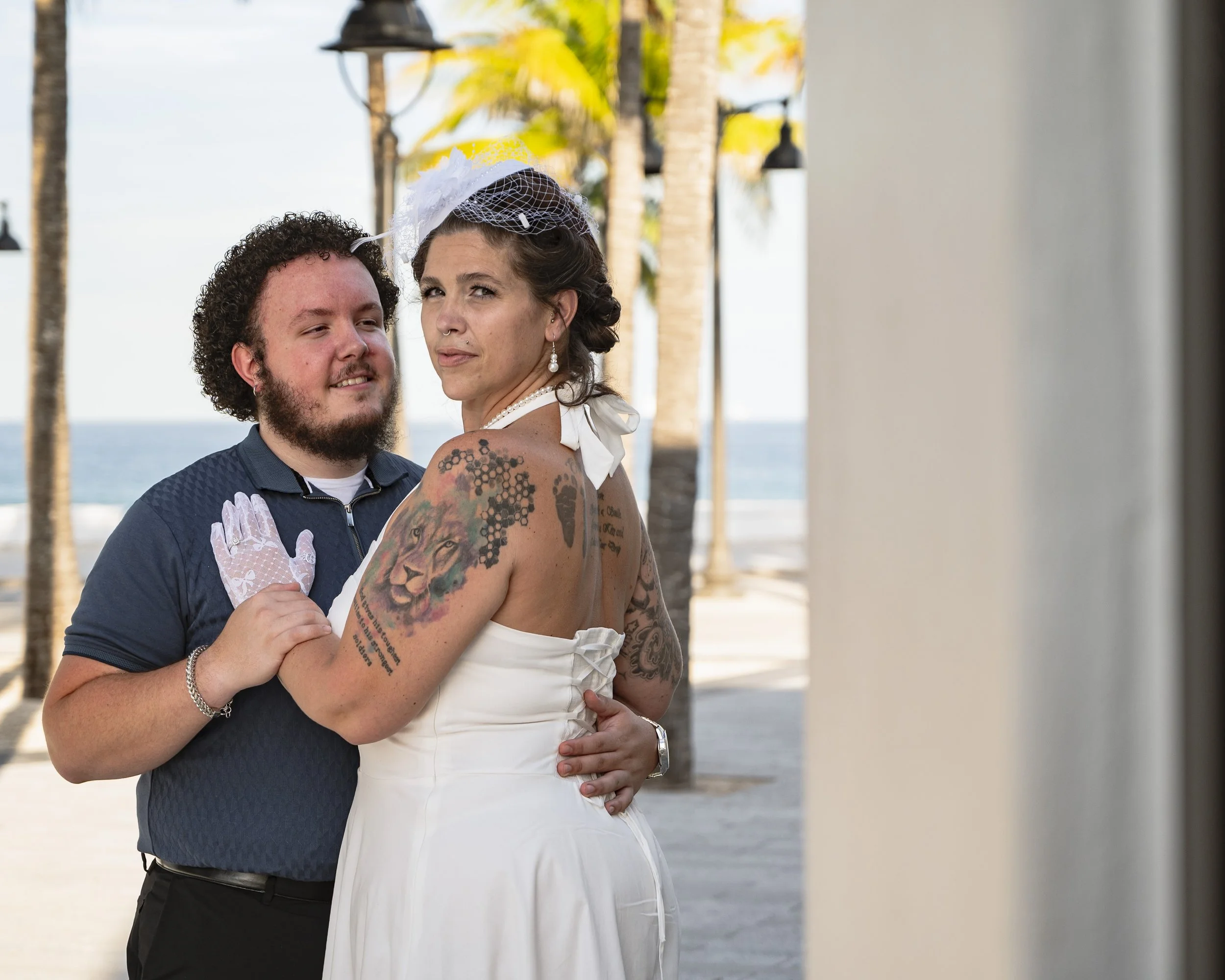 A couple stands close together on a beachside promenade with palm trees and the ocean in the background. The woman is wearing a white dress and a small hat, while the man is wearing a dark polo shirt. They are embracing, with the woman looking at the