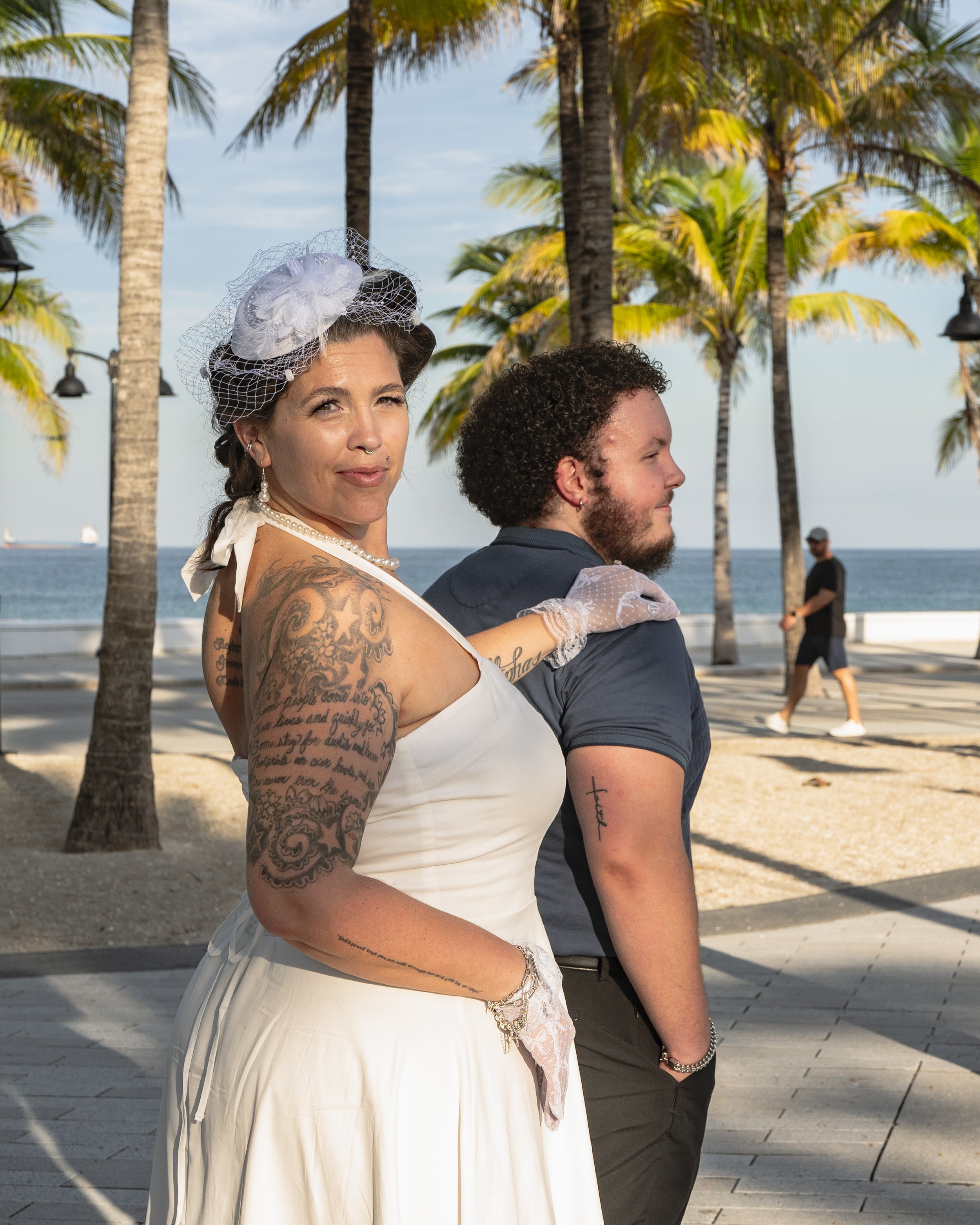A woman with tattoos and vintage attire, wearing a white dress and hat, stands by a man with curly hair and a beard, dressed in a dark t-shirt, near palm trees and the ocean.