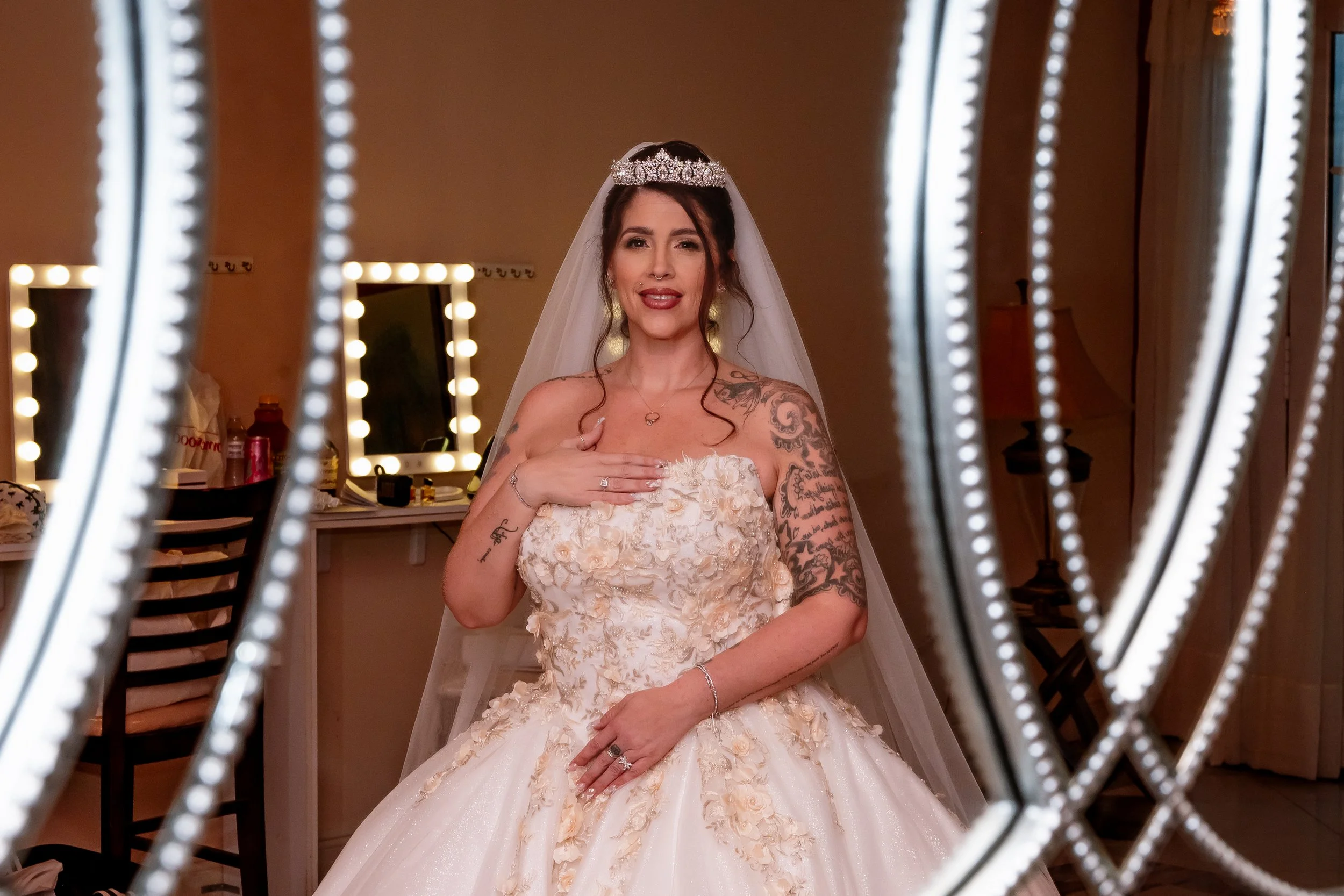 A smiling woman in a wedding dress and tiara, with tattoos on her arms and chest, looking into a mirror framed with lights in a dressing room.