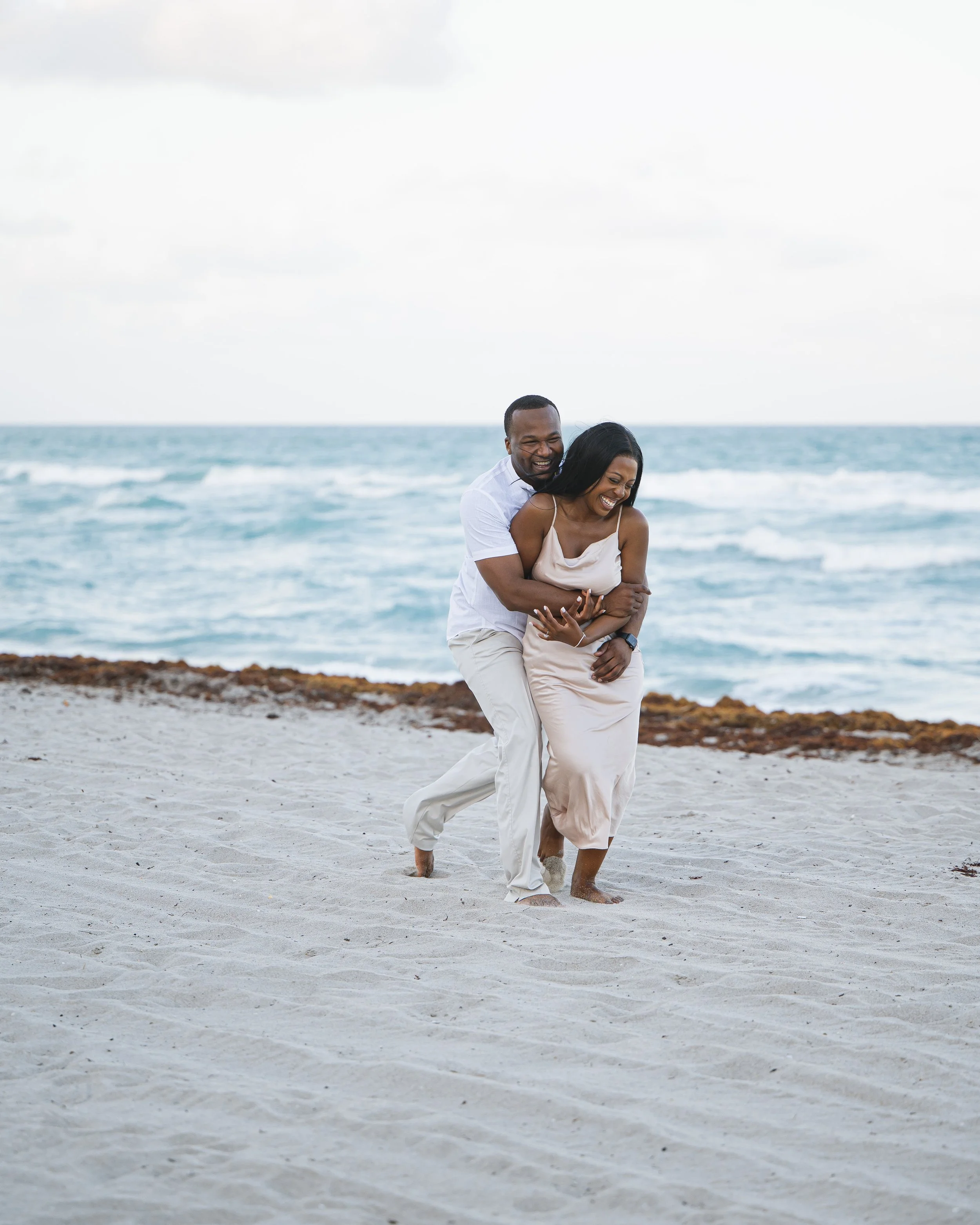 A couple in light-colored clothing playing and laughing on the beach near the ocean.