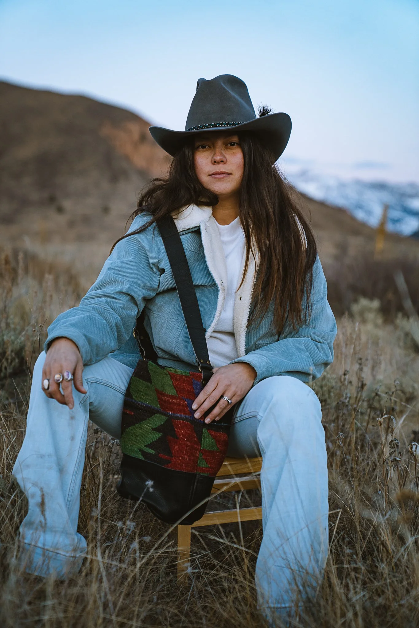 A woman sitting outdoors on a wooden chair in a rural area with dry grass, wearing a cowboy hat, denim jacket, and rings, holding a colorful bag.