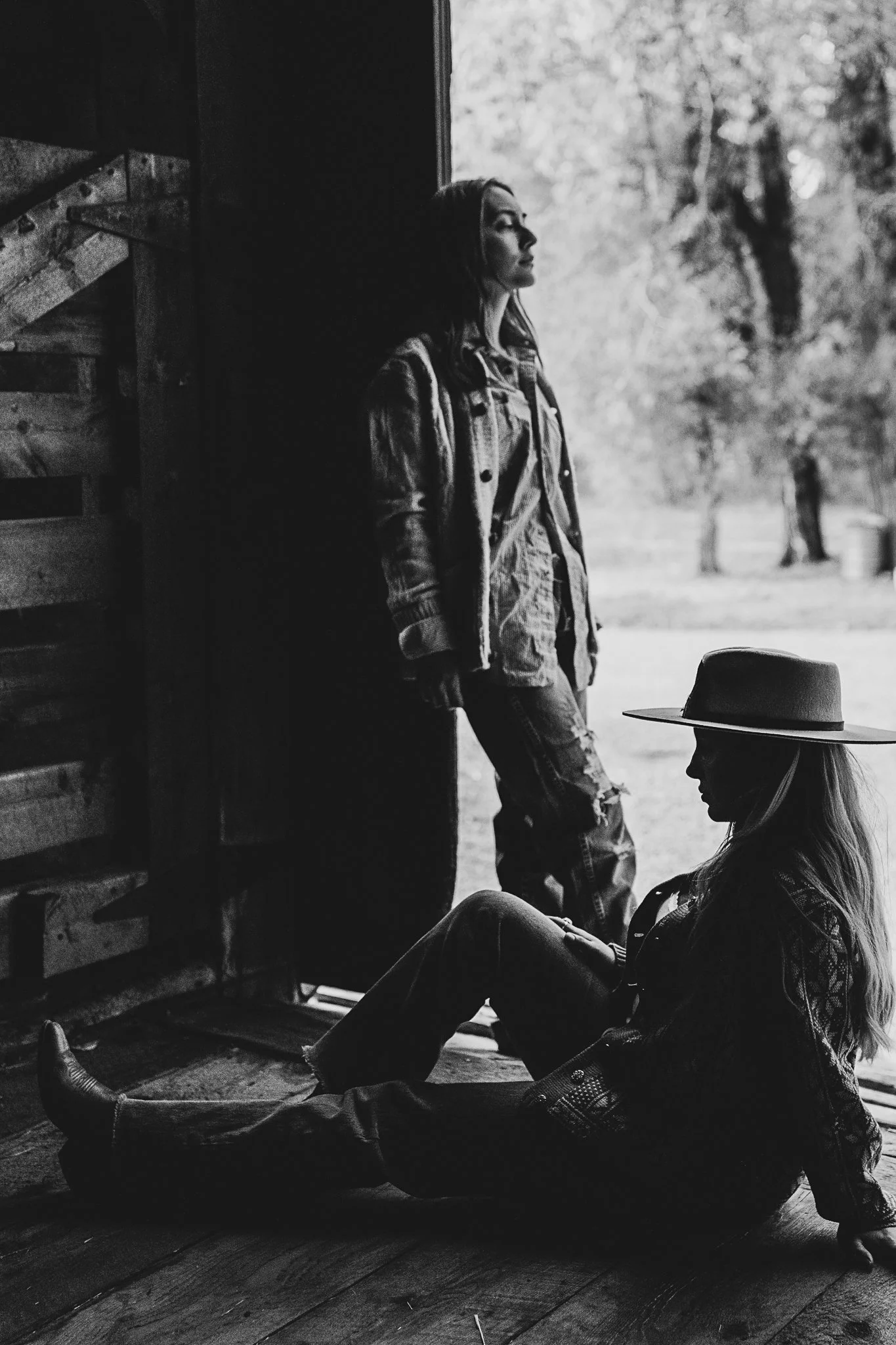 Black and white photo of two women in a rustic wooden barn, one standing and one sitting, with trees outside in the background. The woman sitting wears a wide-brimmed hat.