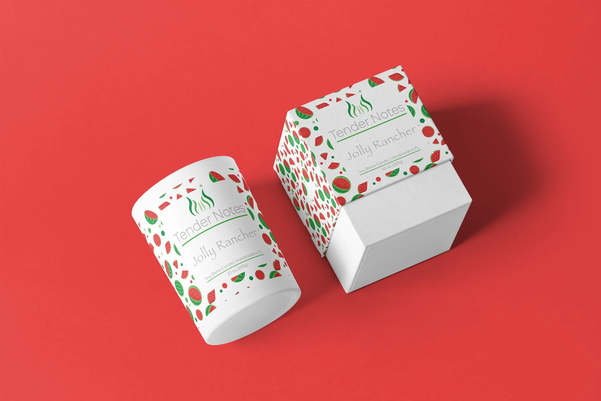 A white candle with a decorative label featuring green and red watermelon and strawberry patterns, labeled 'Tender Notes Jolly Rancher', placed next to a matching box on a coral background.
