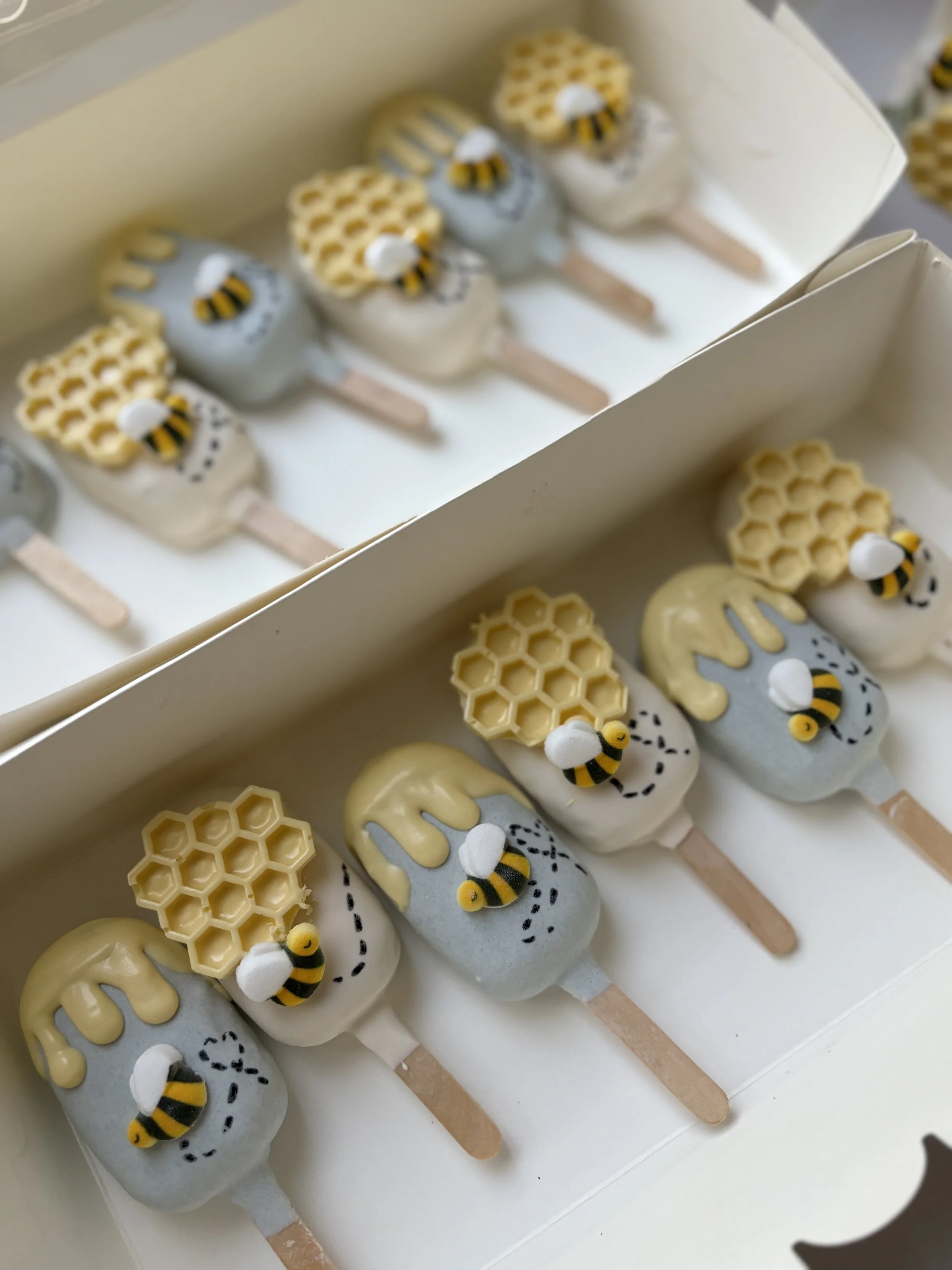 Box of bee-themed cake pops decorated with honeycomb, bee figures, and honey drizzles in yellow, black, and gray colors.