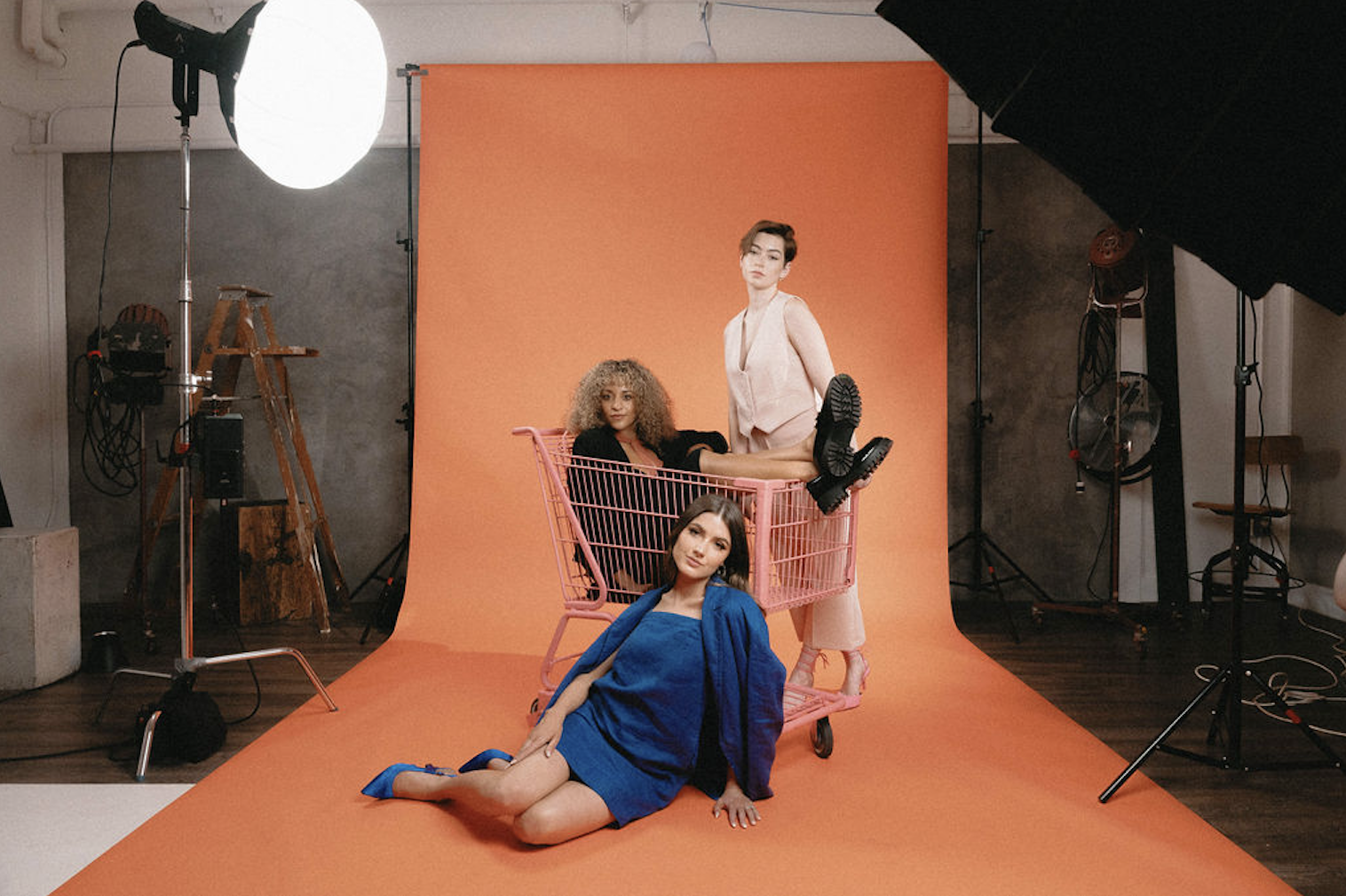Scott Social team members are sitting on an orange background in a photo studio. They are wearing a blue linen monochrome outfit, a light pink matching set, and a black set.