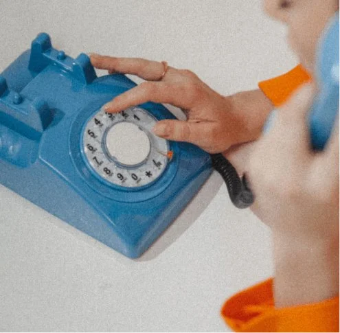 A woman in an orange jacket is slightly off camera, she is dialing with her finger on a rotary telephone that is bright blue.