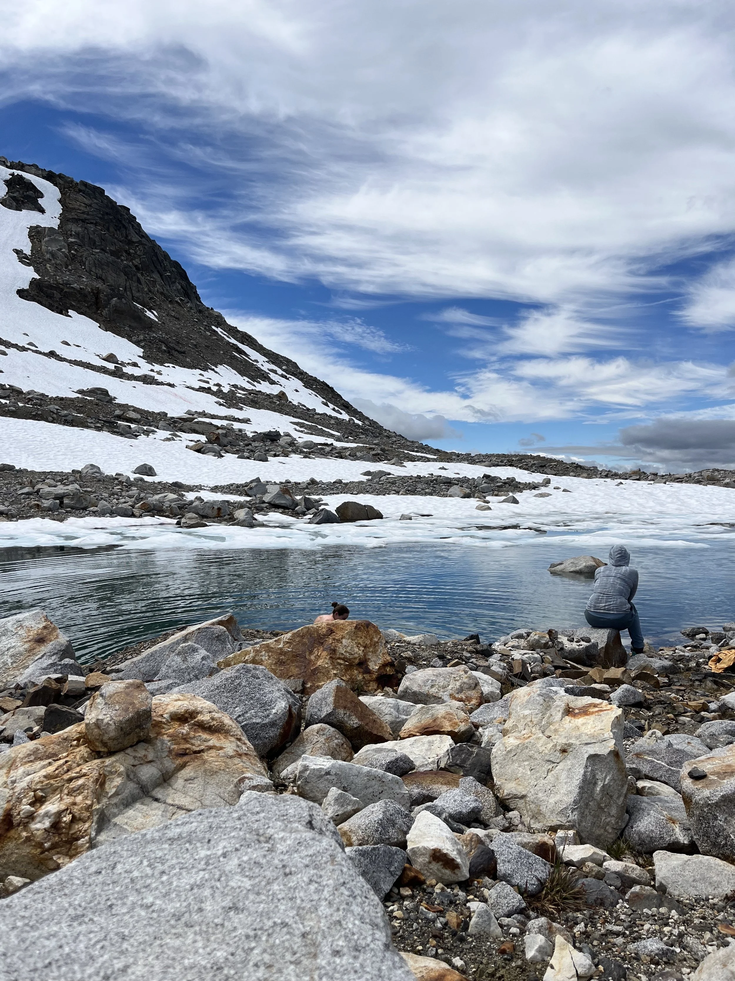 People sitting by a mountain lake with snow-covered landscape and a cloudy sky.