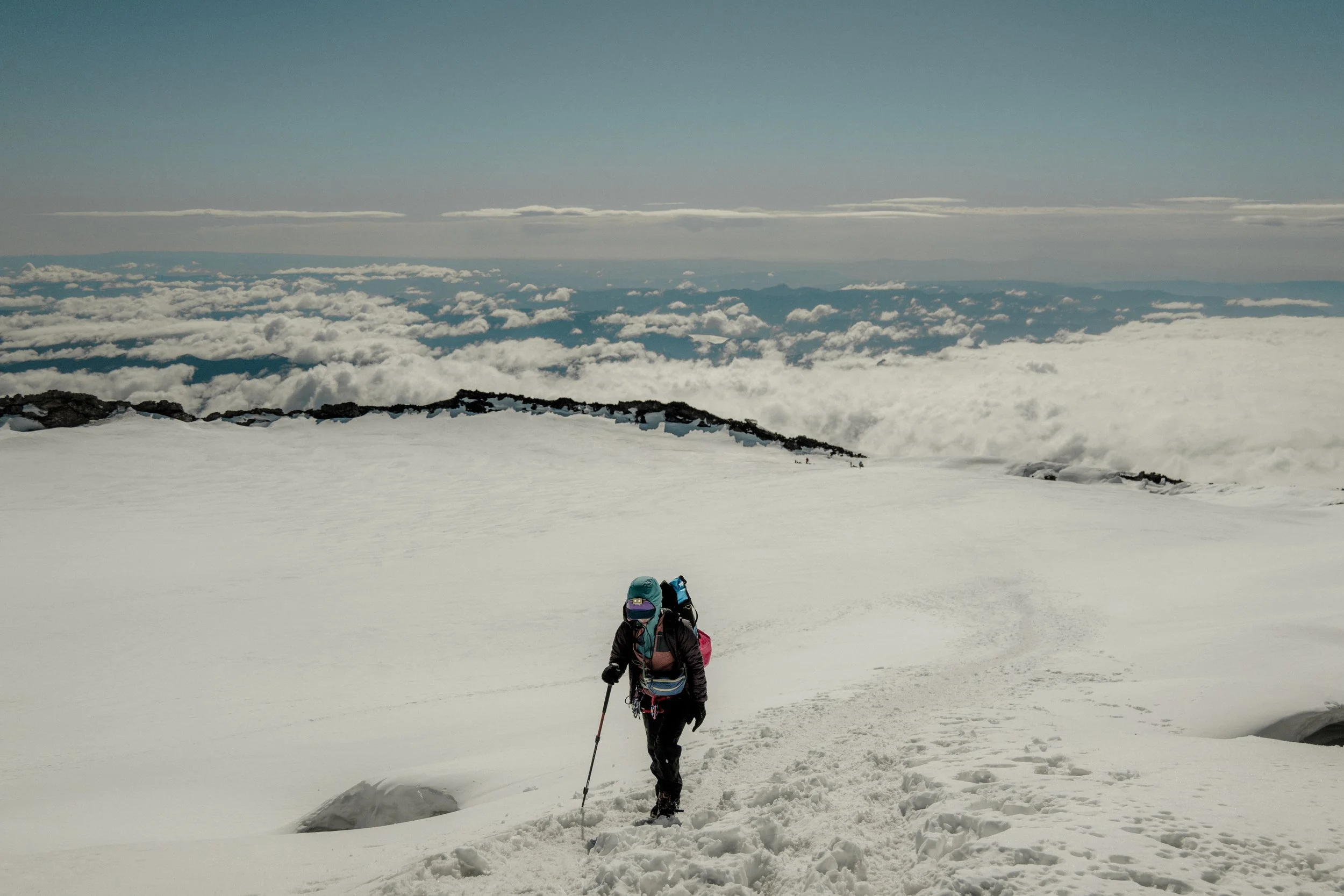 A person hiking in snowy mountain terrain with a backpack, using trekking poles, under a cloudy sky with clouds below the mountain summit.