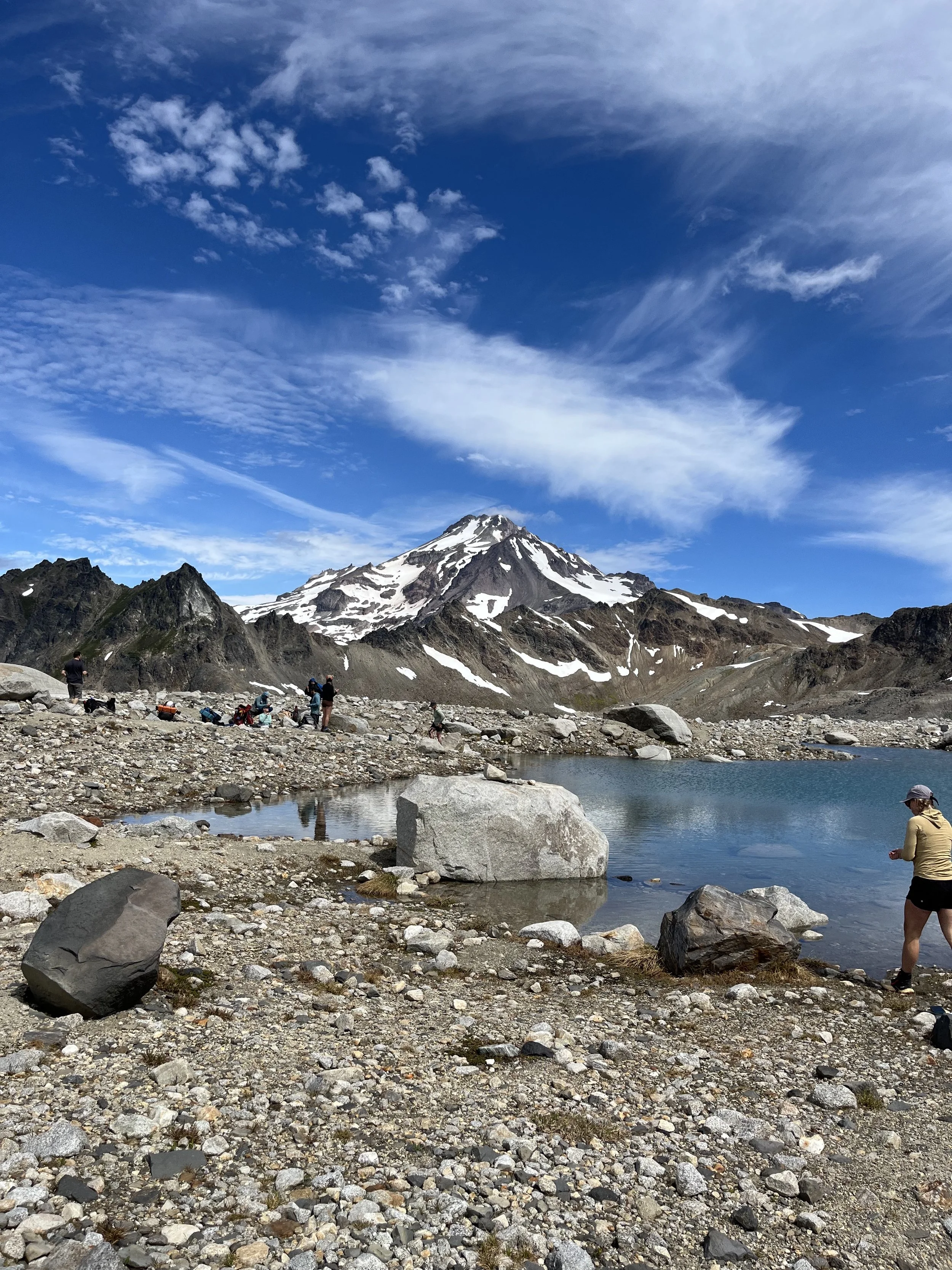 A mountain landscape with a snow-capped peak under a partly cloudy blue sky, with a rocky foreground featuring a small body of water and several people exploring the area.