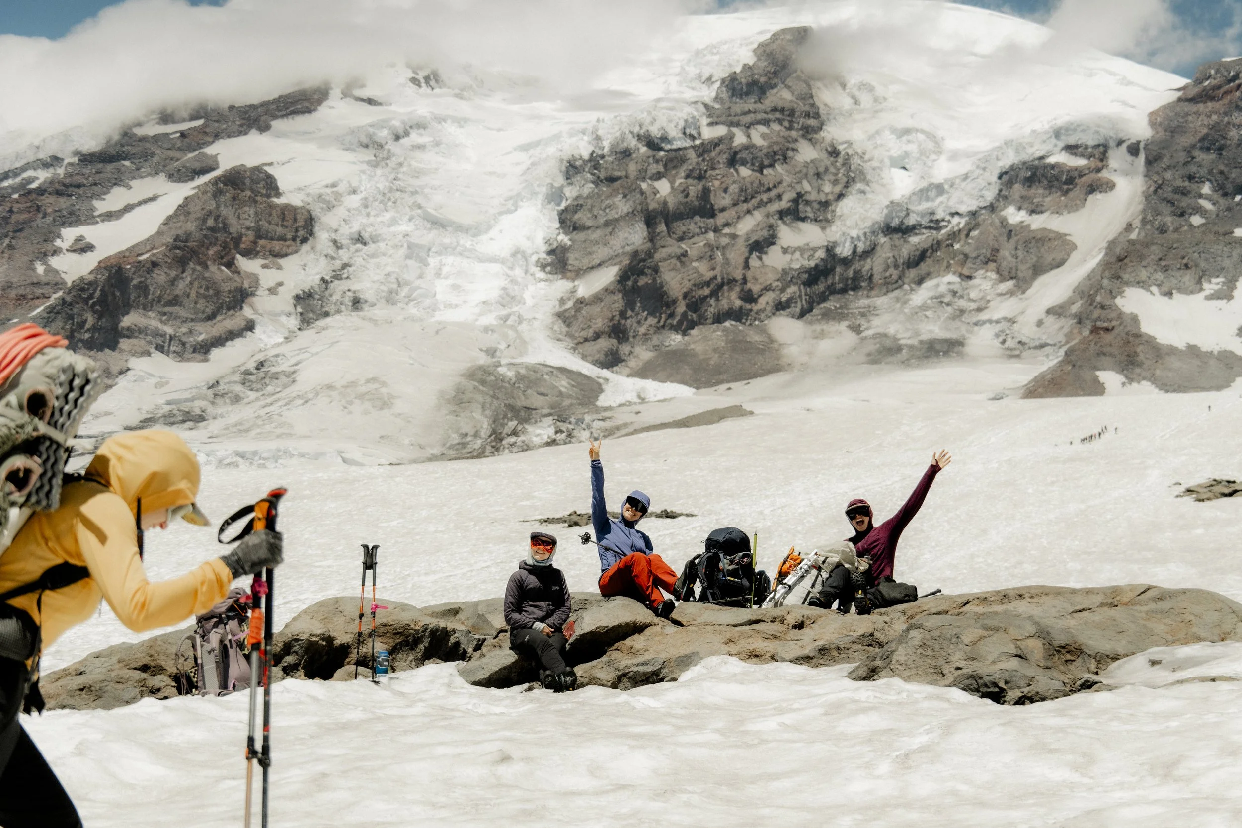 Group of climbers resting and celebrating on a snowy mountain landscape with rocky terrain and glacier in the background.