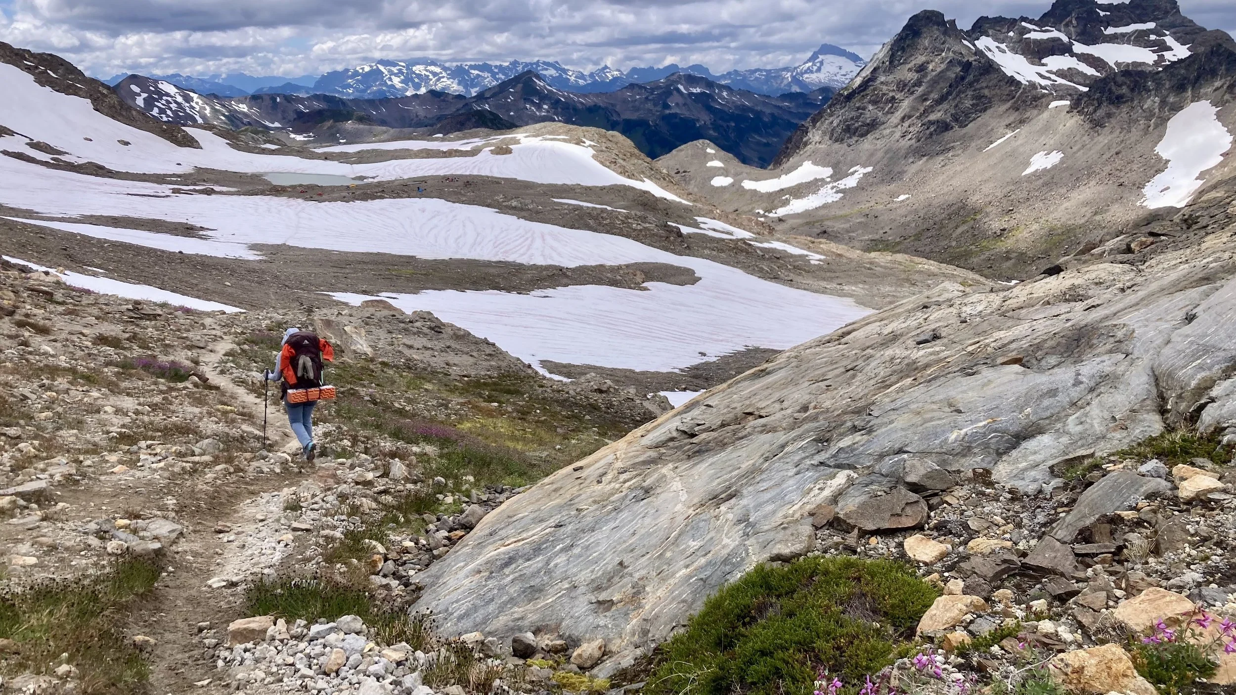A hiker with a backpack walking on a rocky trail in a mountain landscape with snow patches, rugged peaks, and distant mountains.