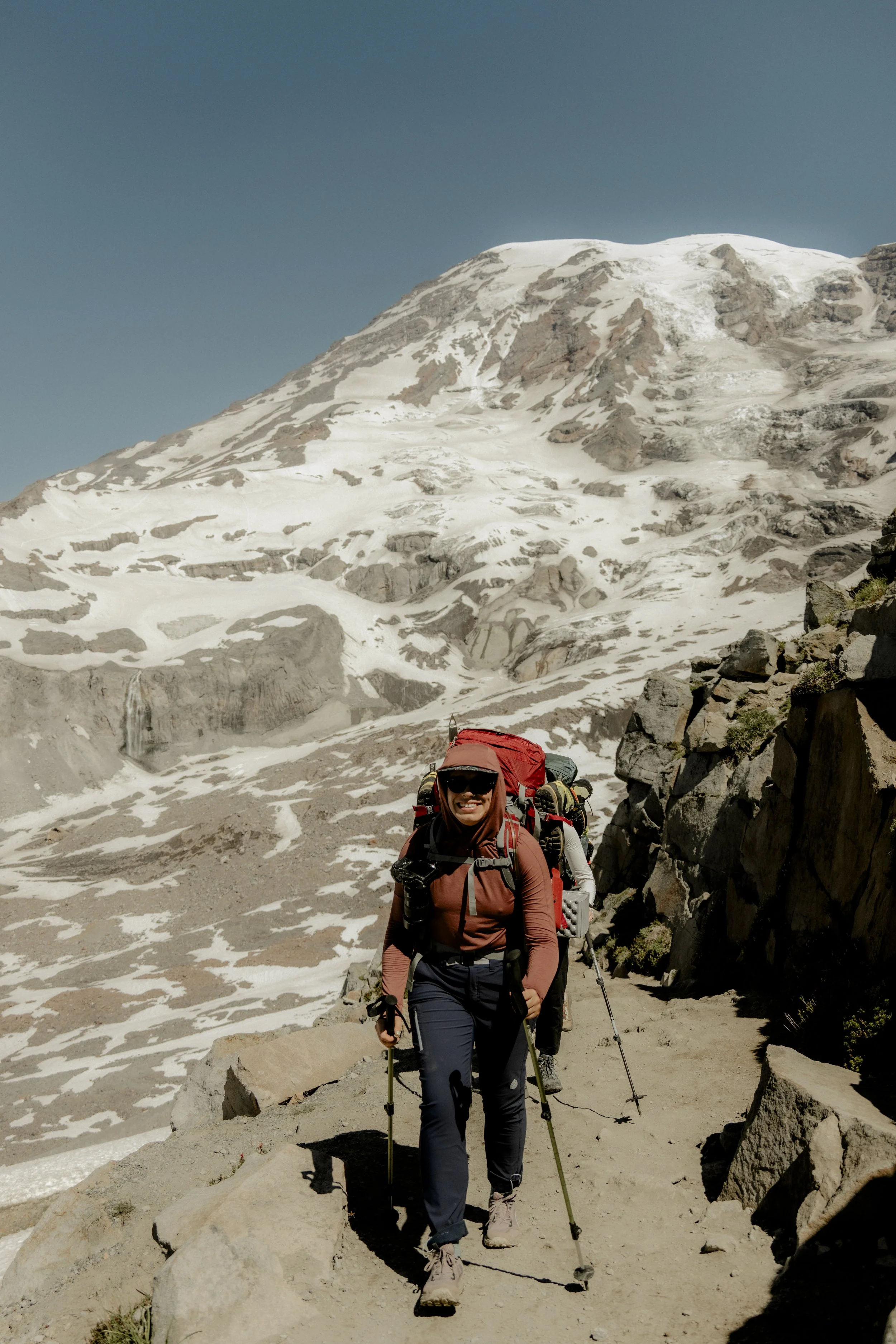 A group of hikers trekking through a mountainous, snow-covered landscape with rocky terrain and a clear blue sky.