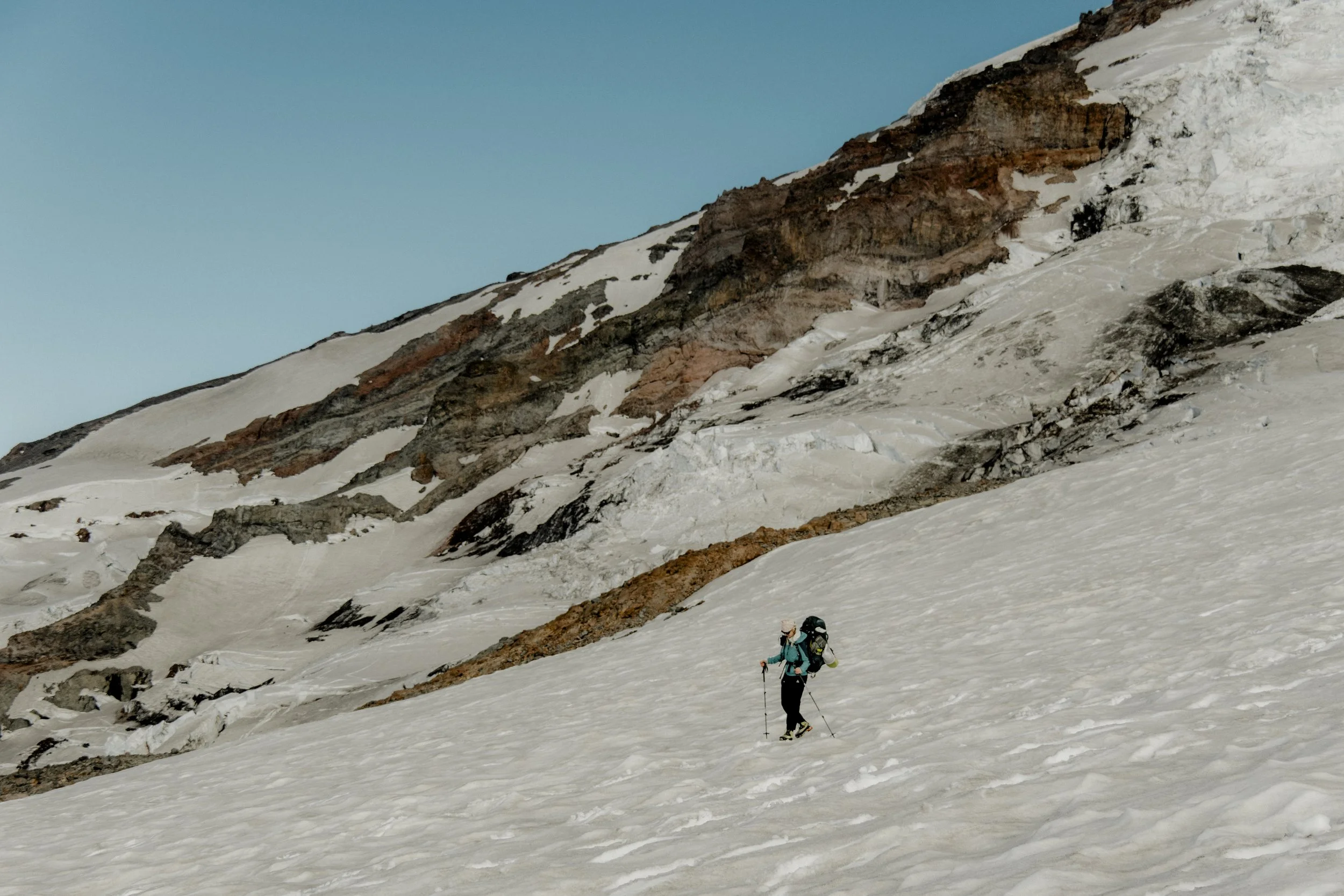 Two skiers trekking across a snow-covered mountain landscape with rocky cliffs and a clear blue sky.