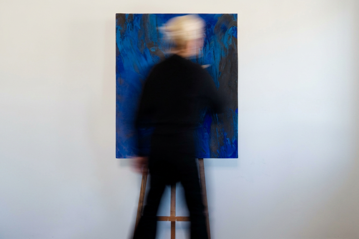 Artist, Sandra Tinari, standing in front of an abstract blue and black painting on a white wall. Portrait of the artist taken in Perth, Western Australia.
