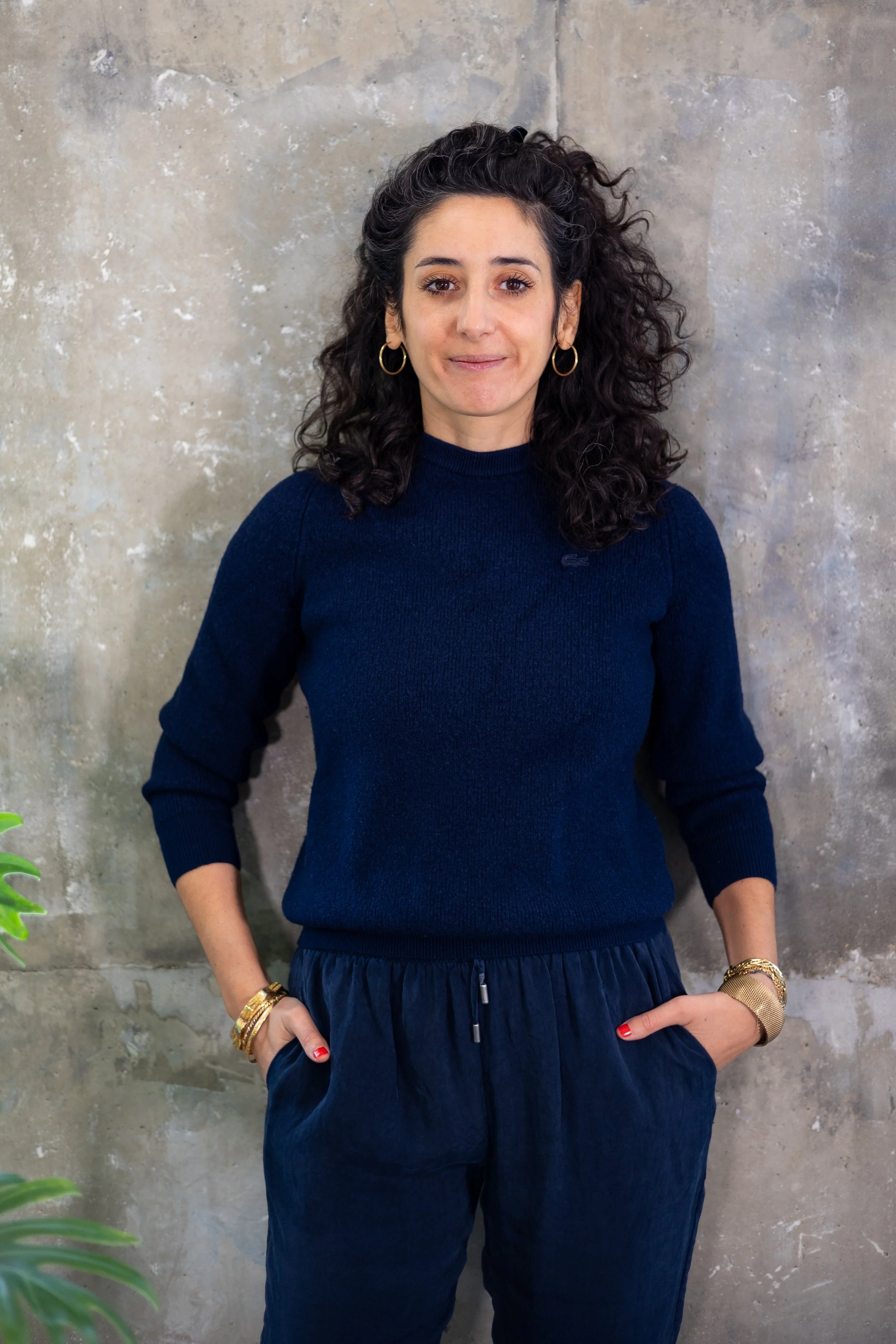 Une femme avec des cheveux bouclés portant un pull bleu foncé, des boucles d'oreilles en cercle et des bracelets dorés, debout devant un mur en béton avec des mains dans les poches.