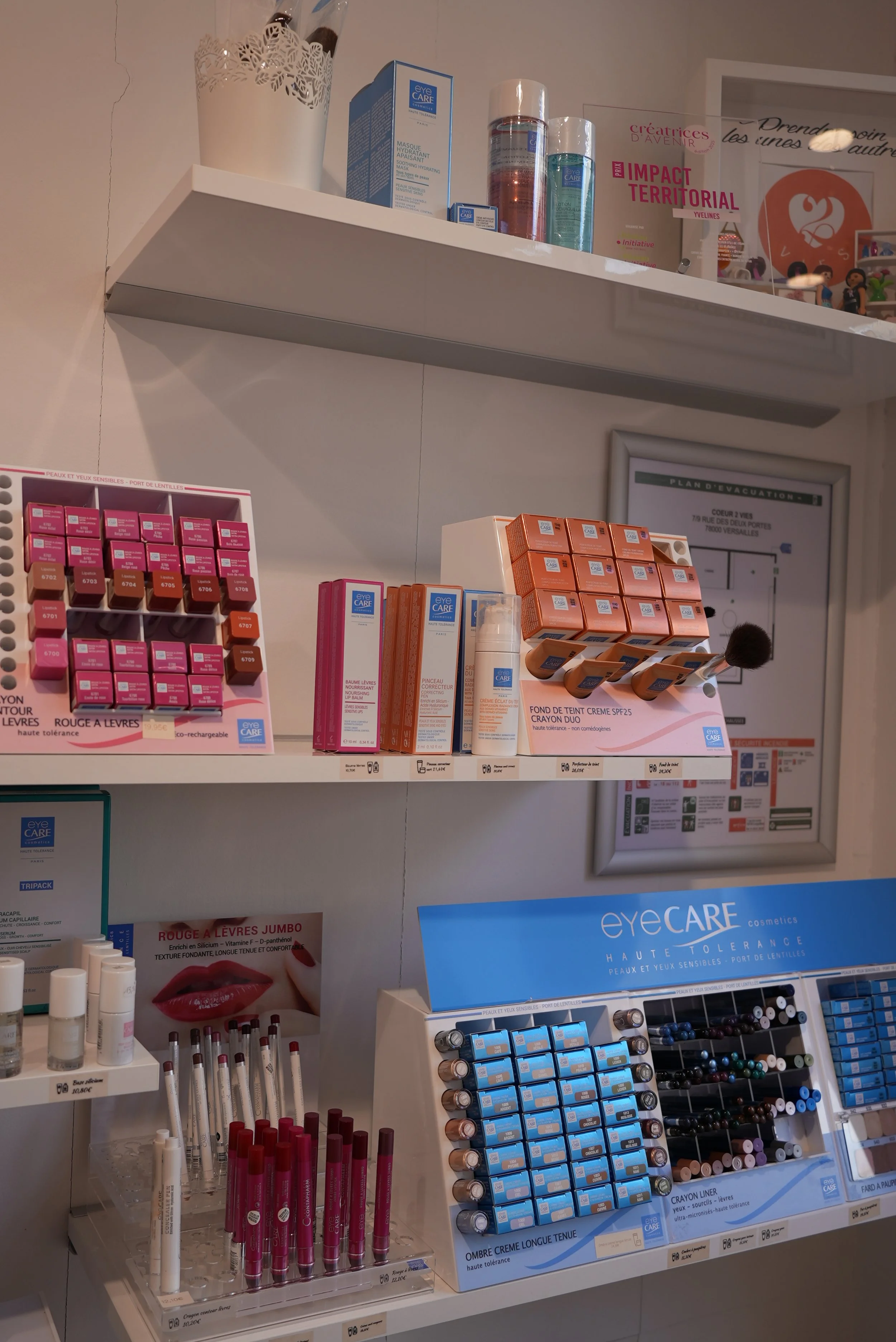 Étagère de produits cosmétiques pour les yeux et les lèvres, notamment des rouges à lèvres, des crayon pour les yeux, et des produits pour l'entretien des lentilles de contact dans un espace de pharmacie ou institut de beauté.