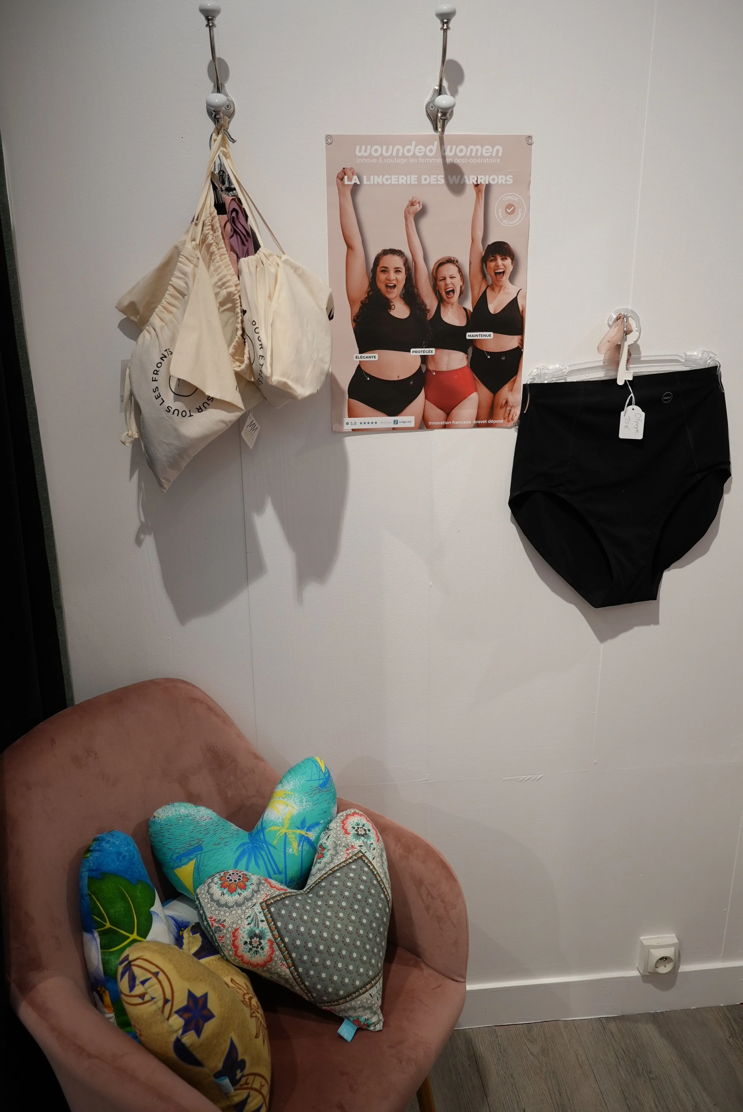 Une chambre avec un fauteuil rose contenant plusieurs coussins colorés et un poster sur le mur représentant trois femmes en sous-vêtements. Deux sacs suspendus au mur et une prise électrique au sol.