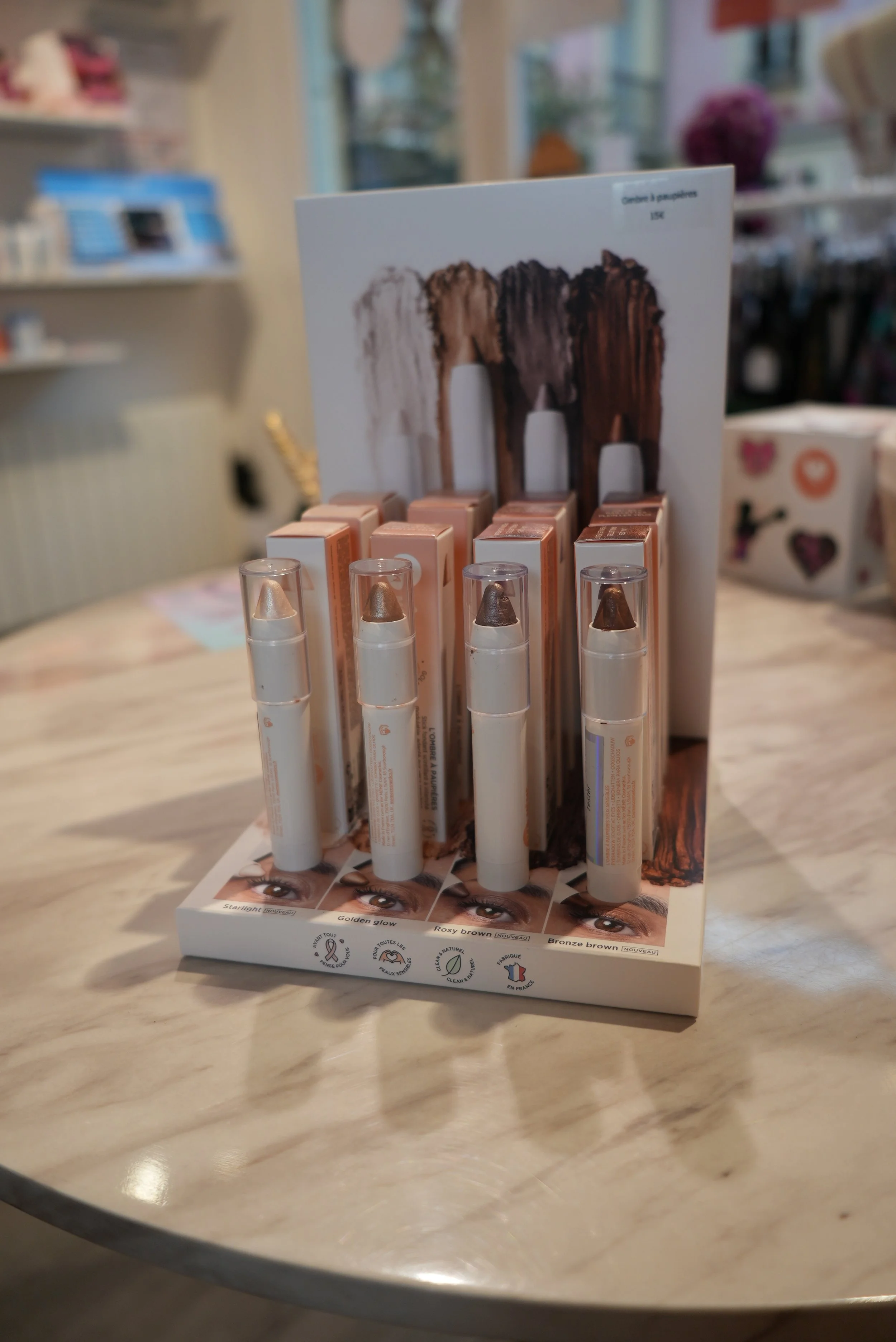 Concealer sticks de différentes teintes sur un présentoir. Le présentoir comporte une affiche avec des images d'yeux et des descriptions des teintes : "Starlight", "Golden glow", "Rosy brown" et "Bronze brown".