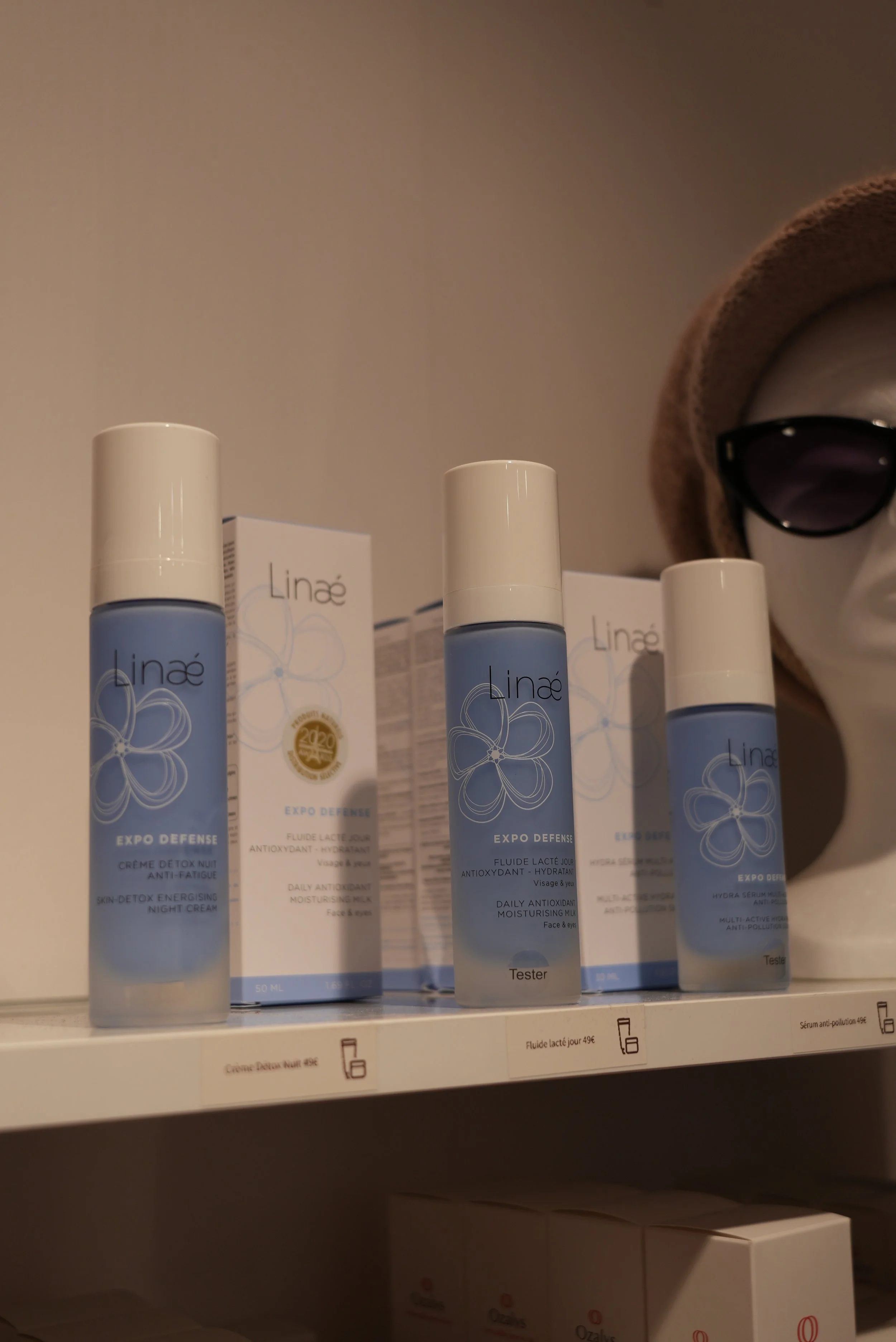 Des bouteilles de soins visage Linæ sur une étagère, avec un masque en forme de visage portant des lunettes de soleil en arrière-plan.