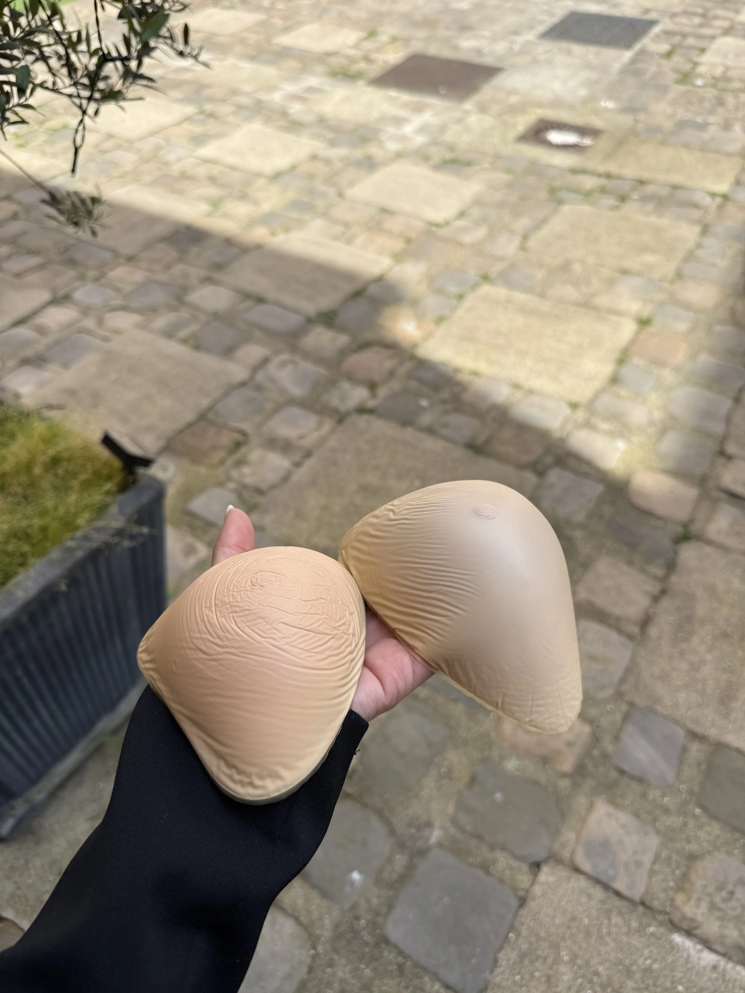 Une personne tient deux cornets de glace beige dans une main, avec un sol pavé en pierre en arrière-plan.
