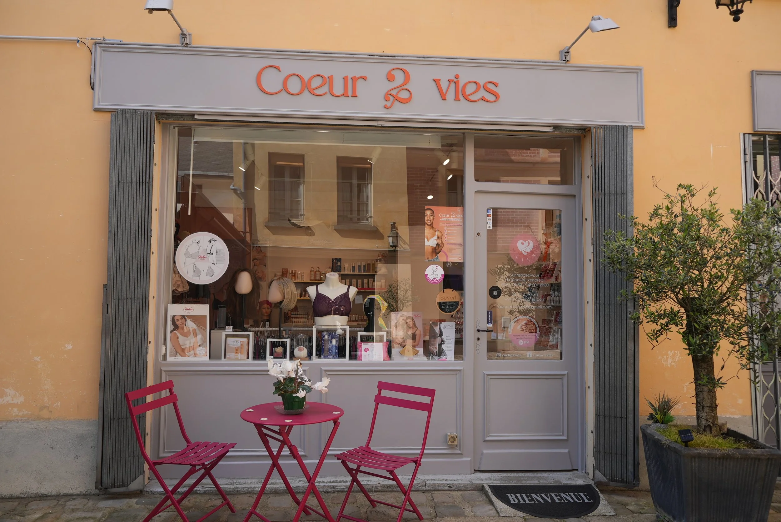Vitrine d'une boutique avec deux mannequins portant des lingerie, affiches promotionnelles, à l'extérieur, il y a une table rouge avec deux chaises et un pot de fleurs, un tapis avec la mention 'BIENVENUE'