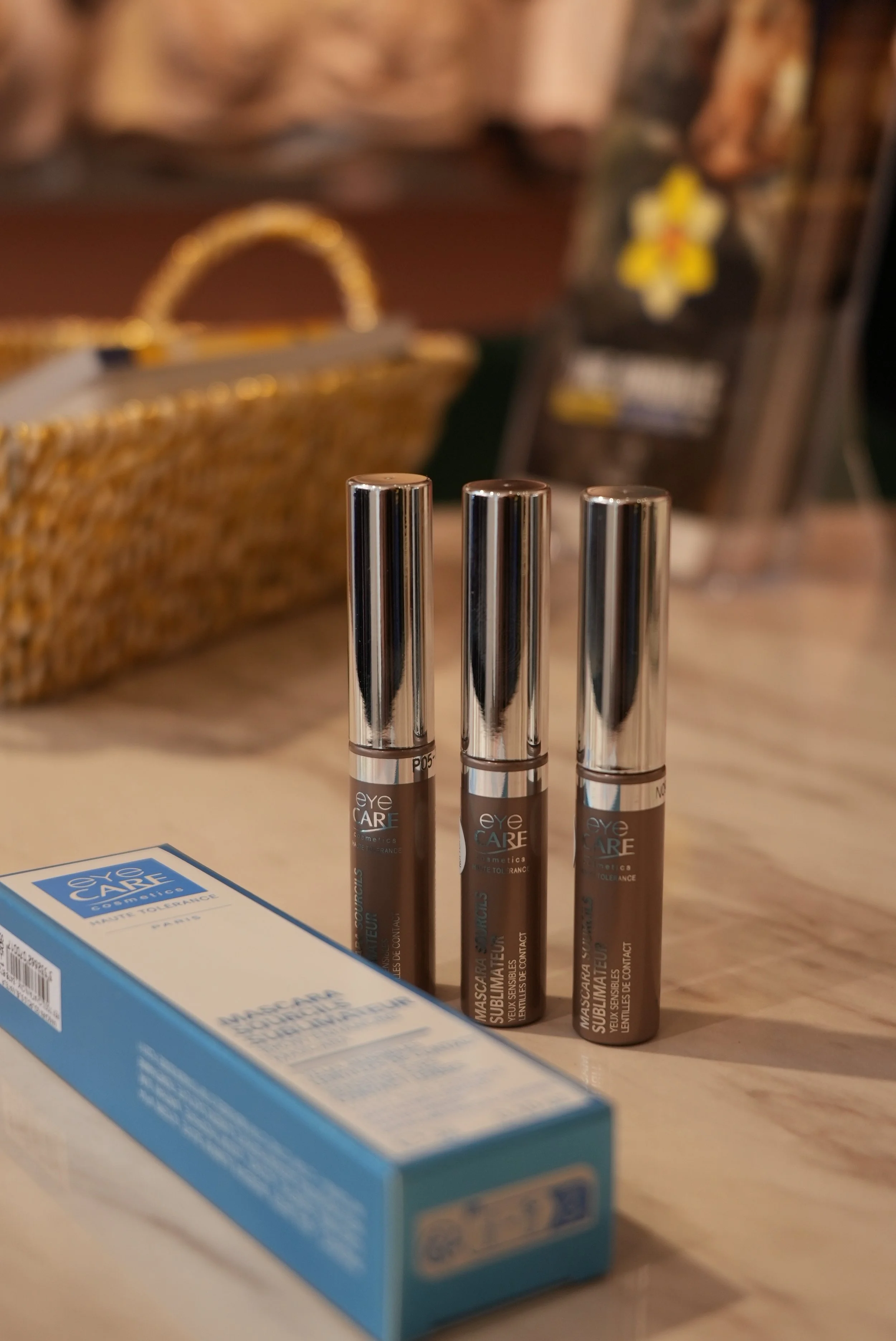 Trois tubes de mascara positionnés devant une boîte bleue, en arrière-plan un panier en osier et des objets décoratifs, dont une fleur en tissu jaune.