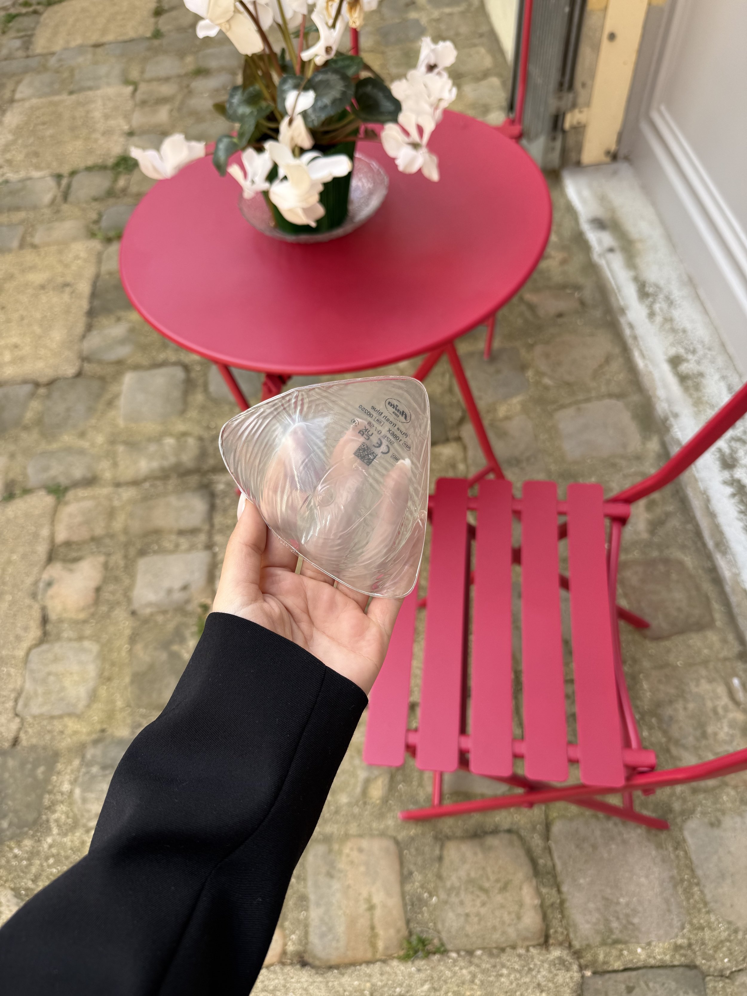 Main dans une manche noire tenant un bol en plastique transparent, devant une table rose et une chaise rose à l'extérieur sur un sol en pierre. Une vase avec des fleurs blanches est placée sur la table.