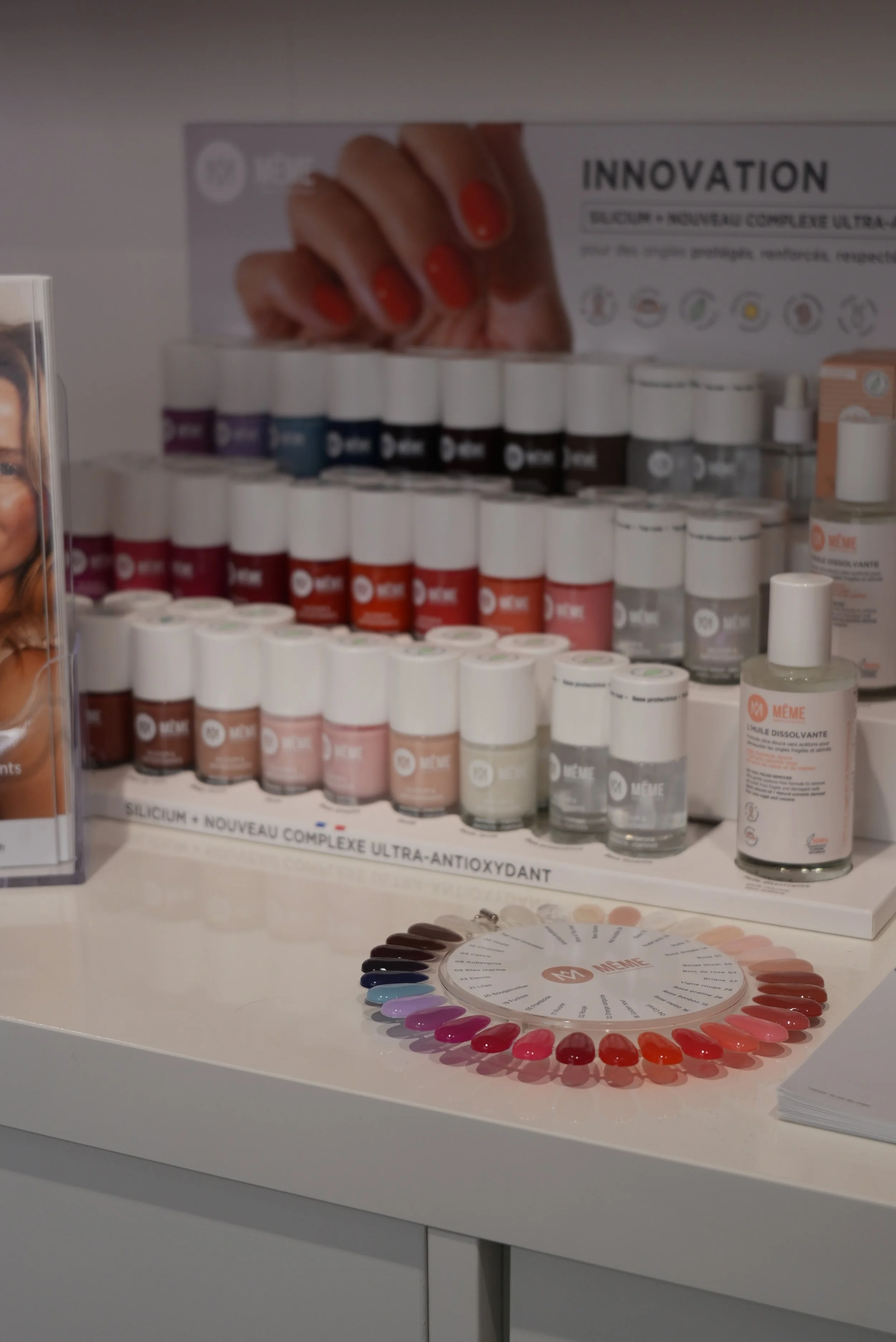 Échantillon de vernis à ongles de différentes couleurs disposé en cercle sur un tapis à l'avant d'une display de produits de soins pour les ongles, avec une affiche promotionnelle en arrière-plan.