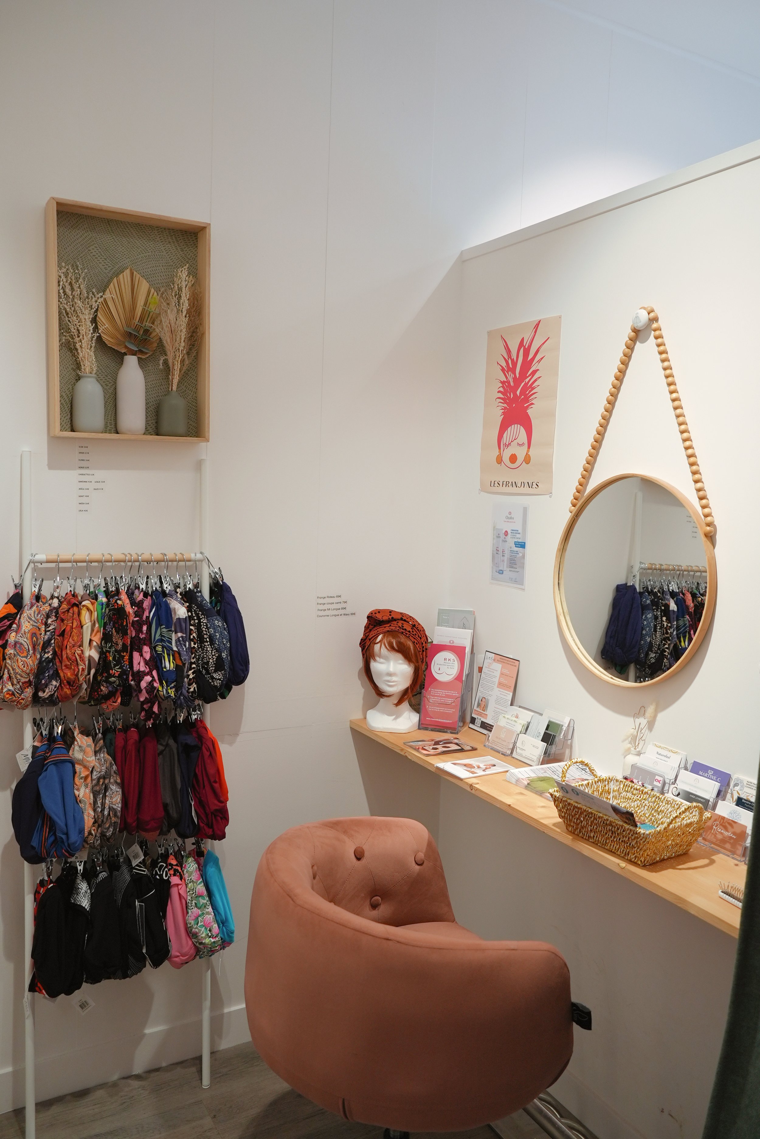 Espace boutique avec un mur d'étagères contenant des vases décoratifs avec des feuillages et des affiches artistiques, un miroir rond suspendu avec un cadre en bois et une chaîne de perles, une table avec des brochures, un panier en osier et une chaise rose en velours.
