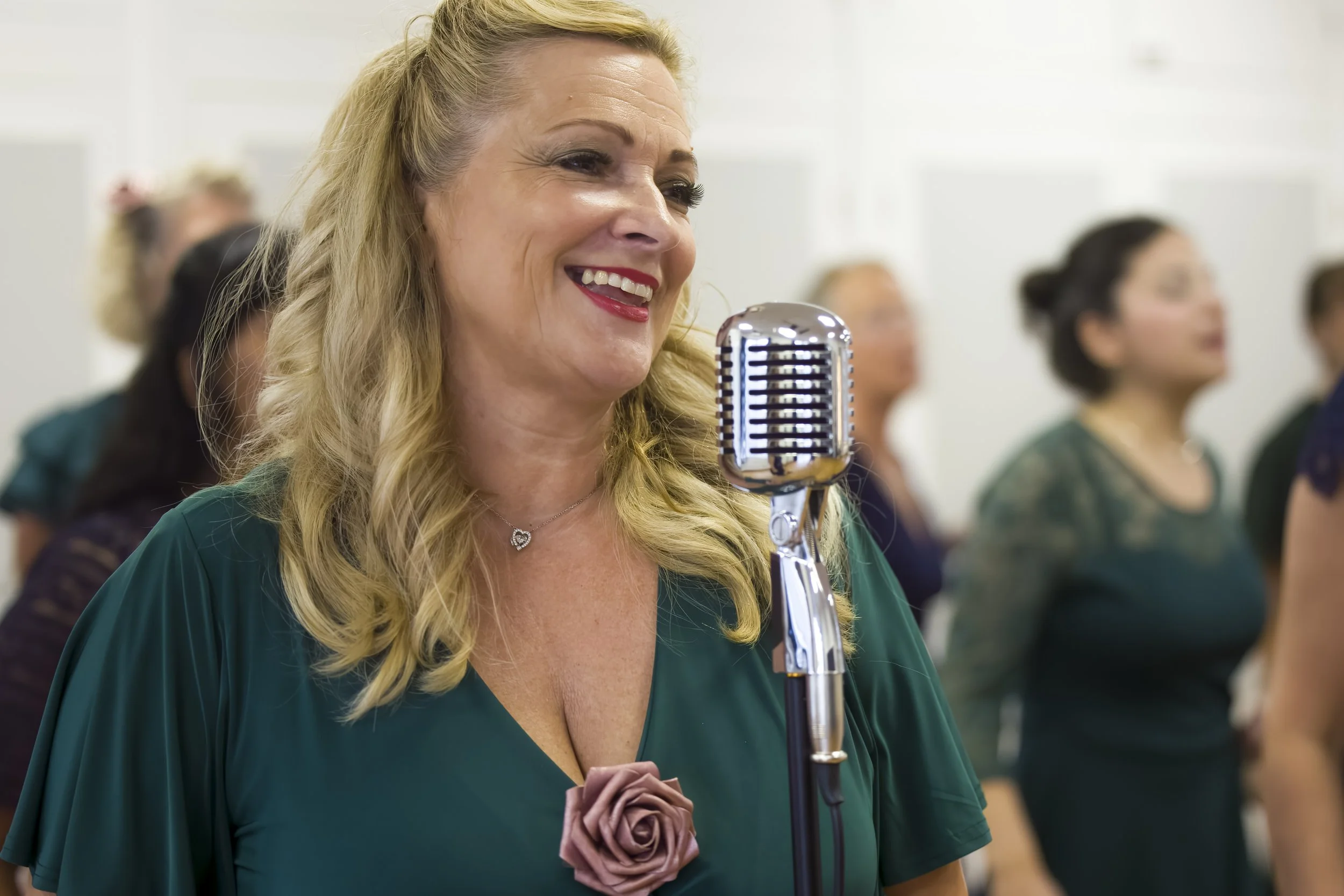 A woman from The Fifty choir with blonde hair smiling while singing into a vintage microphone during a performance in Southend-on-Sea, with several choir members behind her.